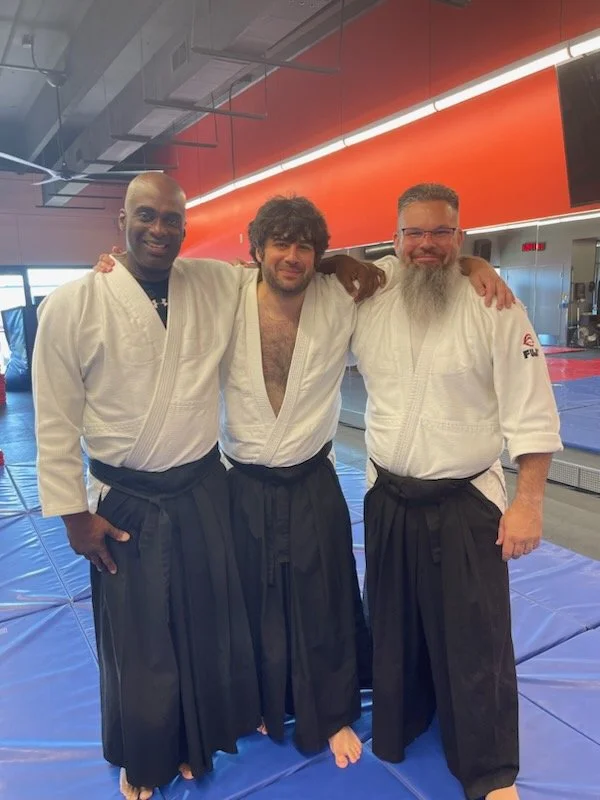 Three men in martial arts uniforms stand together on a training mat in a dojo, smiling with arms around each other.