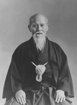 An elderly man with a long beard dressed in traditional Japanese clothing, sitting with hands resting on his lap, against a plain background.