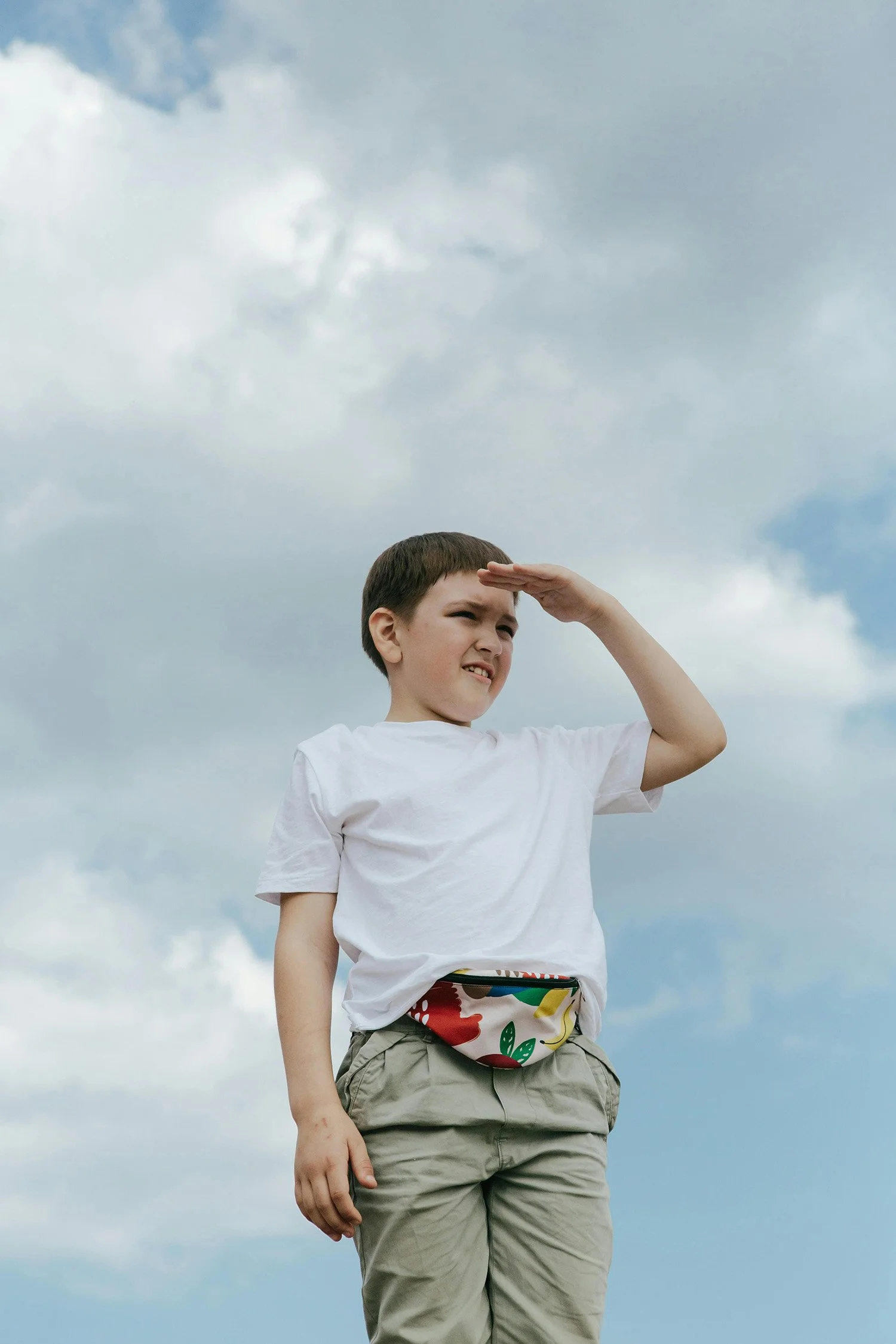 A young boy in a white t-shirt and khaki pants looks into the distance, shielding his eyes with his hand from the sun under a cloudy sky.