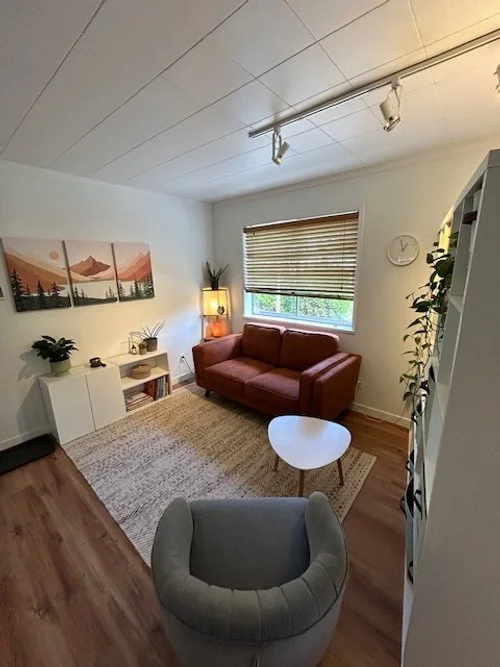 Cozy office with a red sofa, a white coffee table, a grey armchair, a beige rug, and a window with wooden blinds. Wall art and plants decorate the space.
