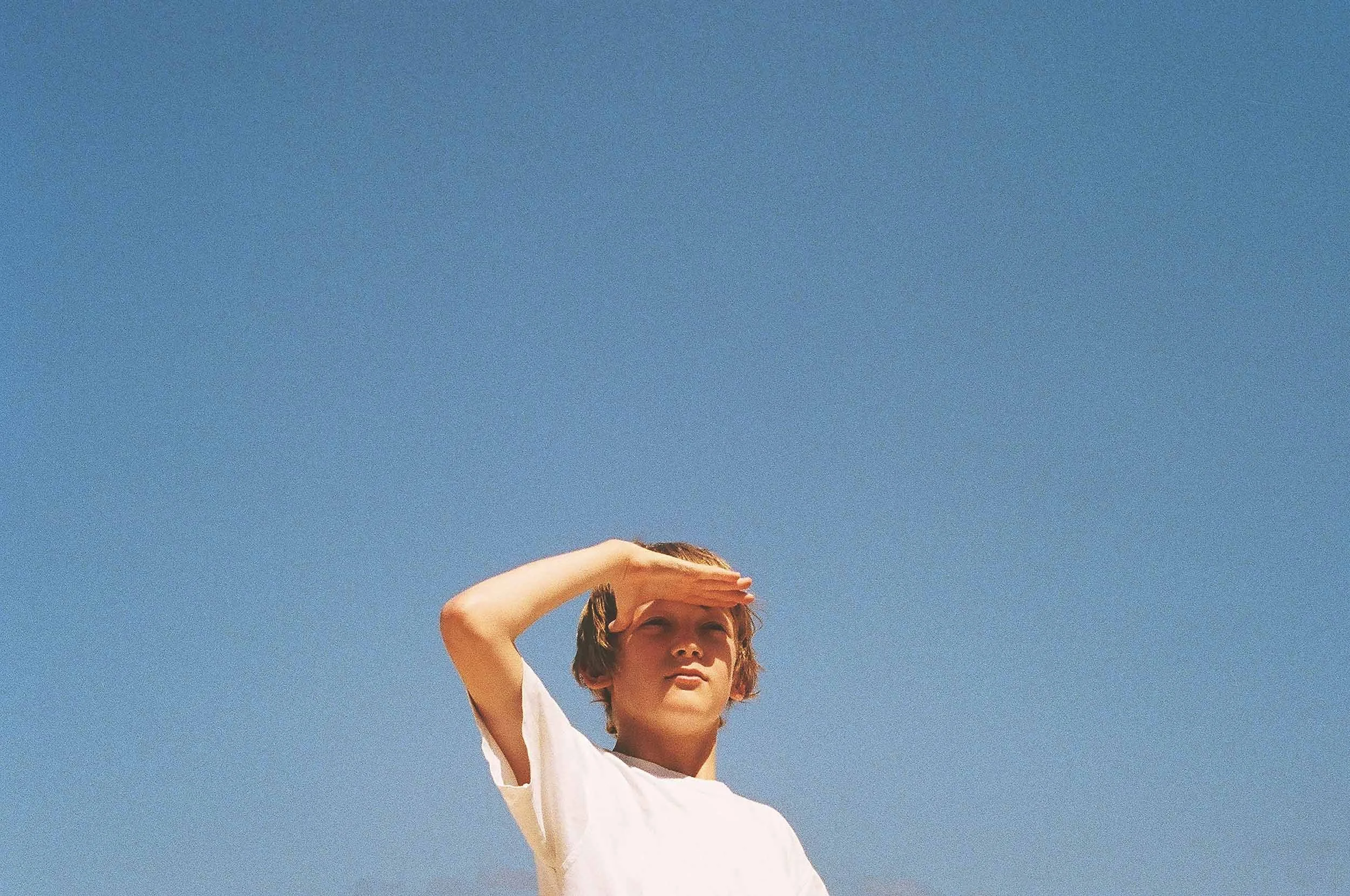 Young boy in a white T-shirt shielding his eyes from the sun with his hand, standing against a clear blue sky.