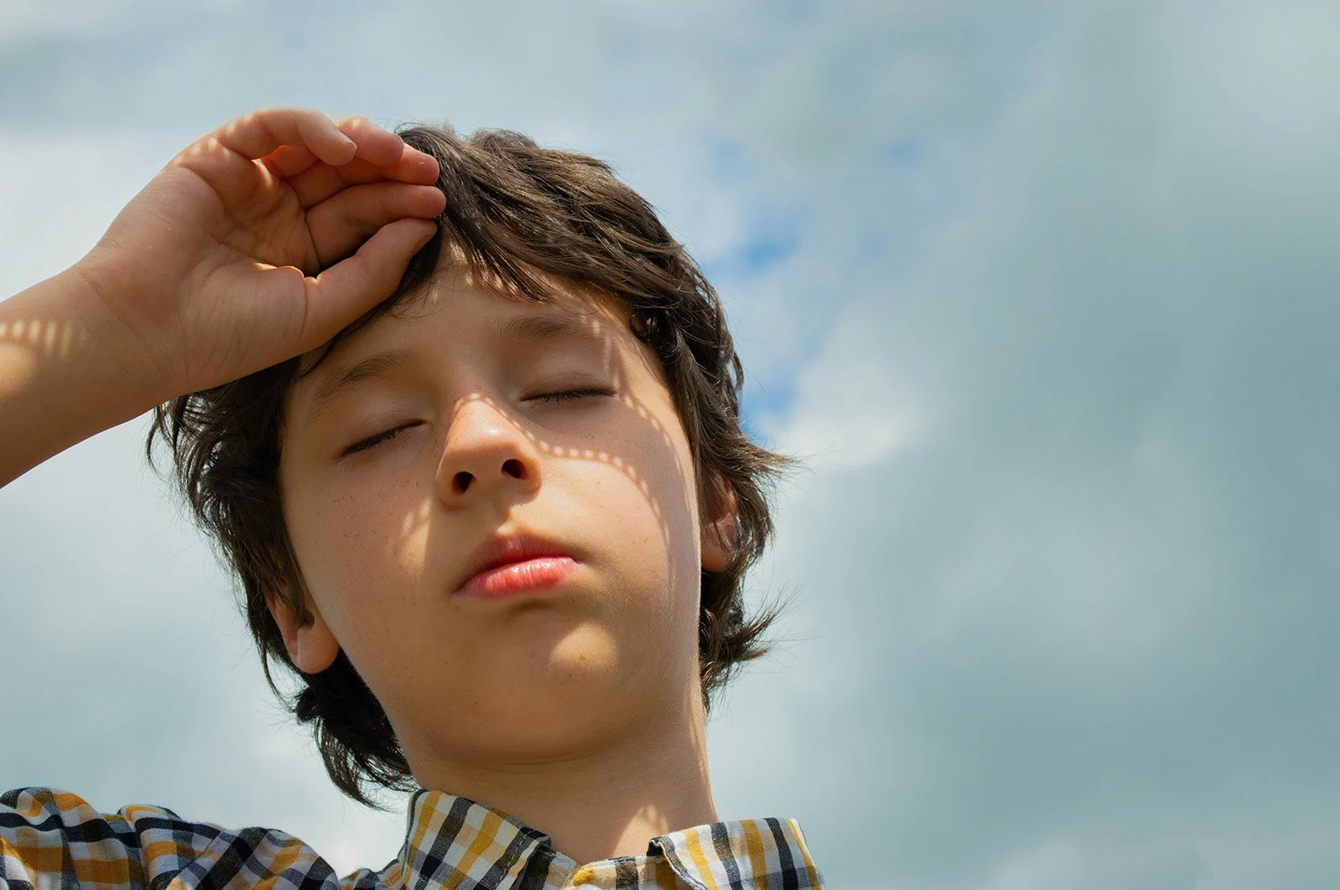 A young boy with closed eyes standing outdoors under a cloudy sky, shading his face from the sun with his hand.