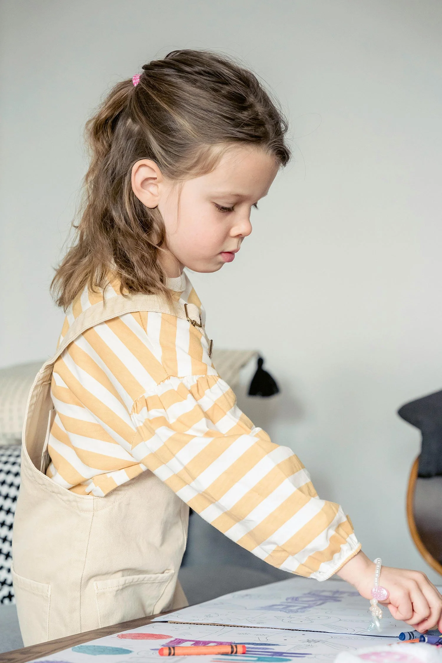 A young girl with brown wavy hair, wearing a yellow and white striped shirt and beige overalls, drawing with colored markers on a large sheet of paper at a table.
