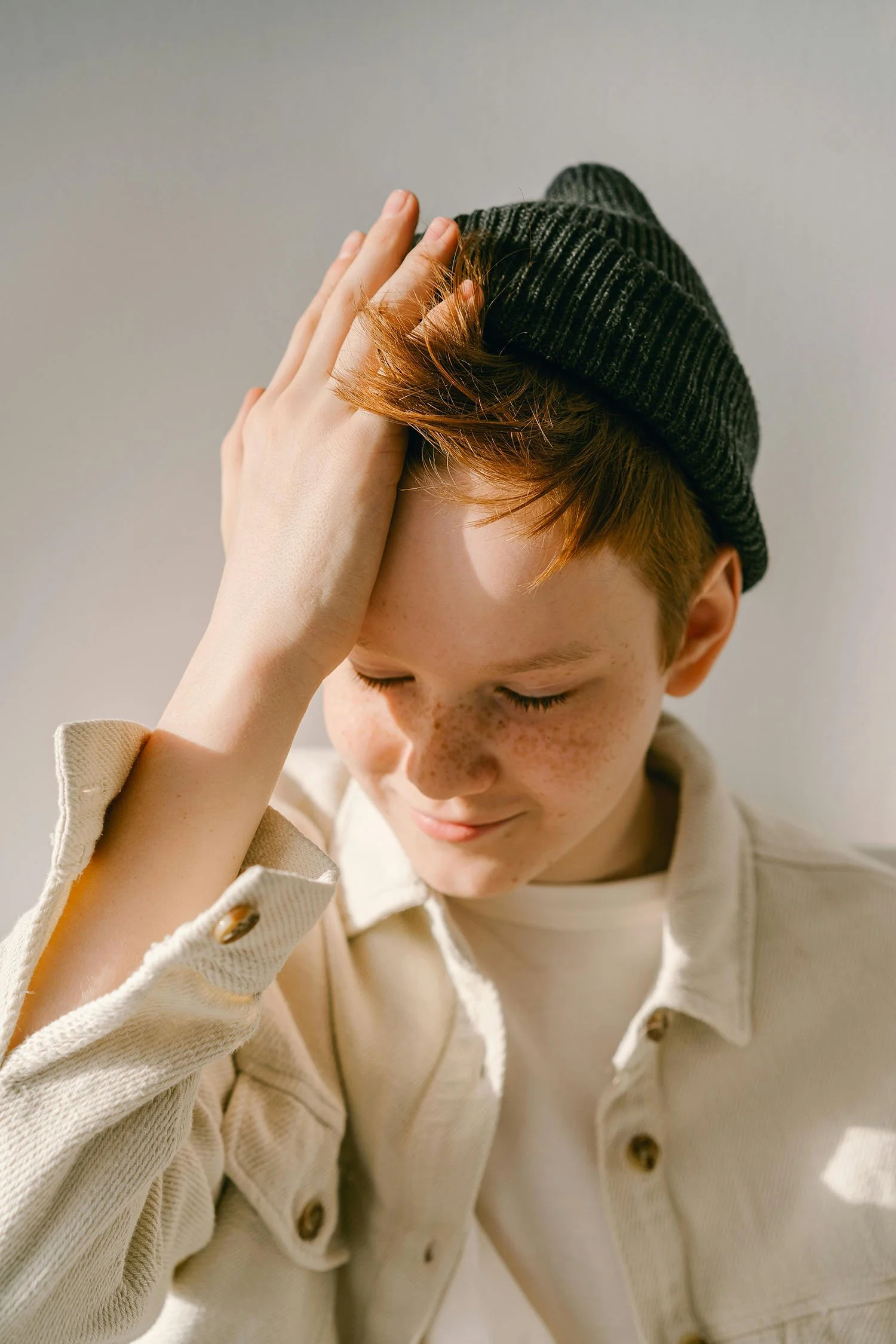 A young boy with red hair and freckles wearing a dark beanie and beige jacket, gently touching his head with his eyes closed and a slight smile.