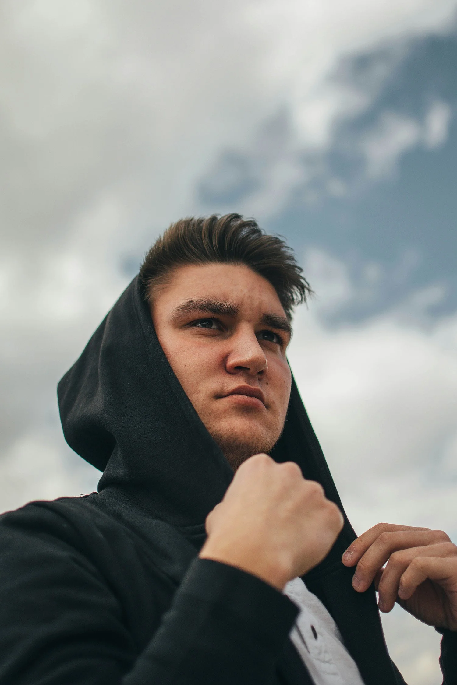 Young man wearing a black hoodie adjusting the hood on his head outdoors with a cloudy sky in the background.