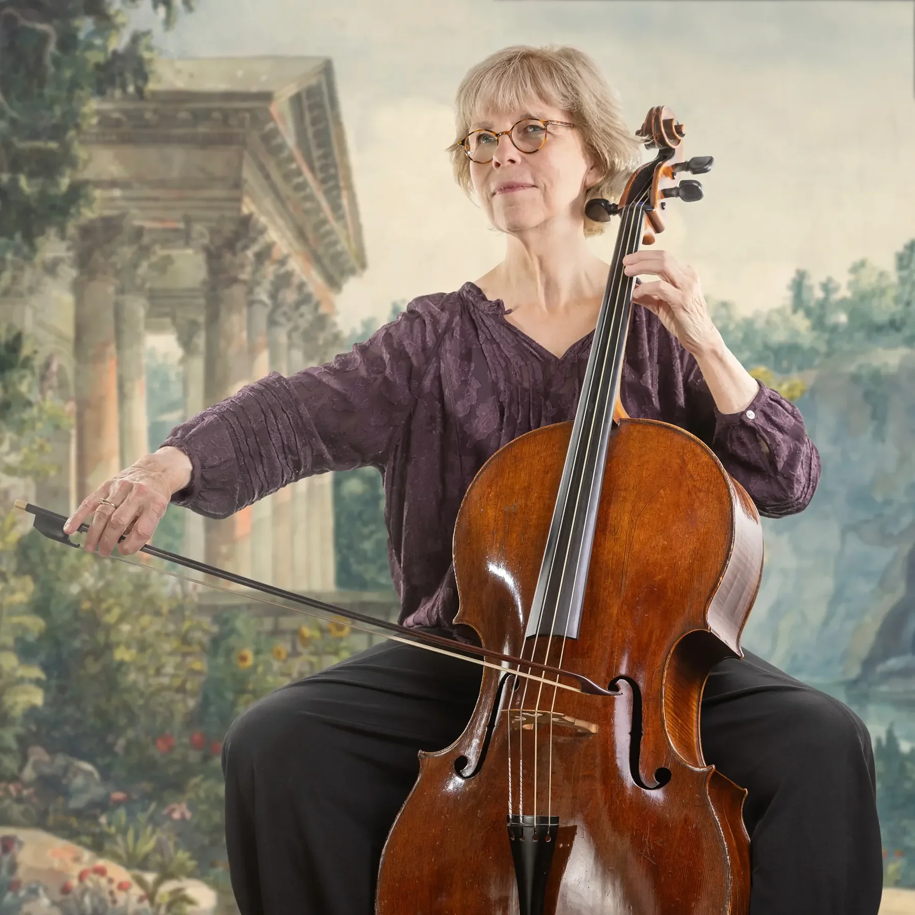 A woman playing a cello in front of a painted backdrop of ancient ruins and greenery.