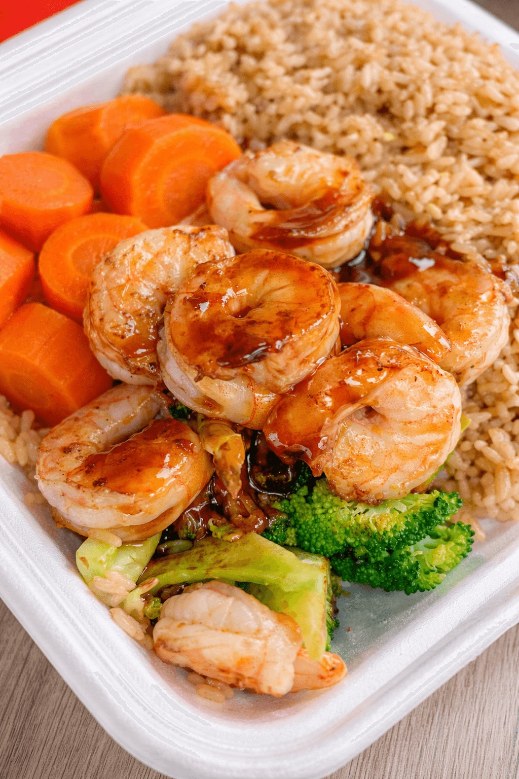 Shrimp with sauce, cooked carrots, broccoli, rice, and other vegetables in a takeout container.