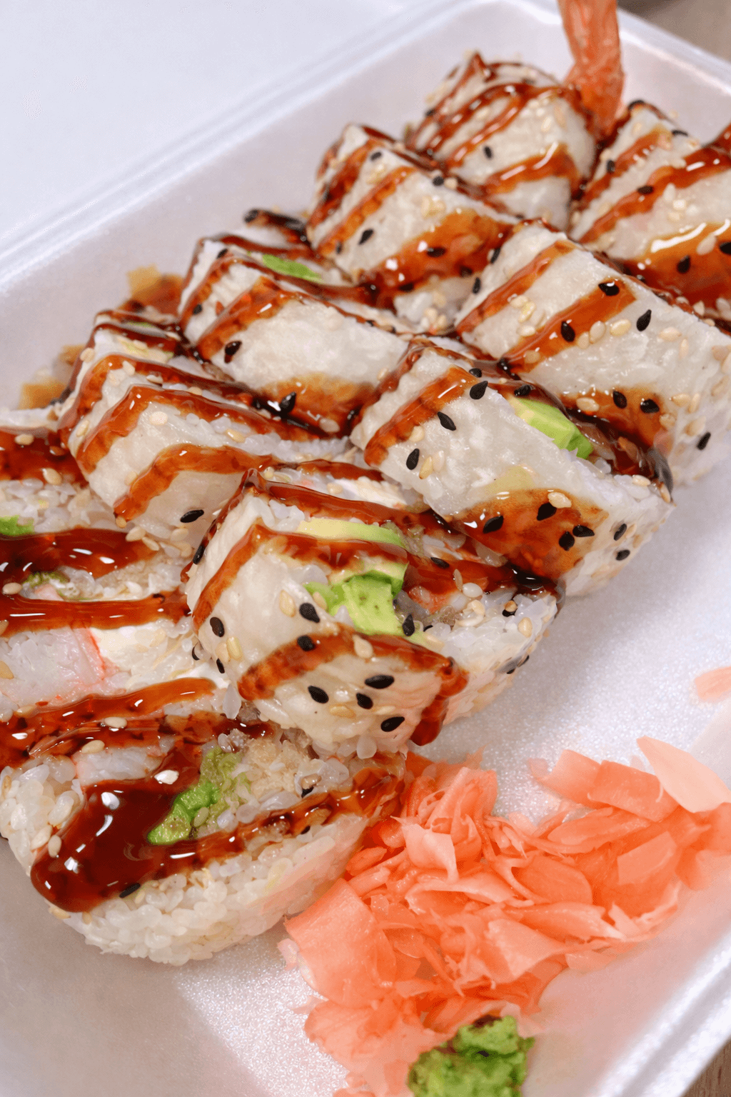 A box of sushi rolls topped with sauce, sesame seeds, and black sesame seeds, with pickled ginger and wasabi on the side.