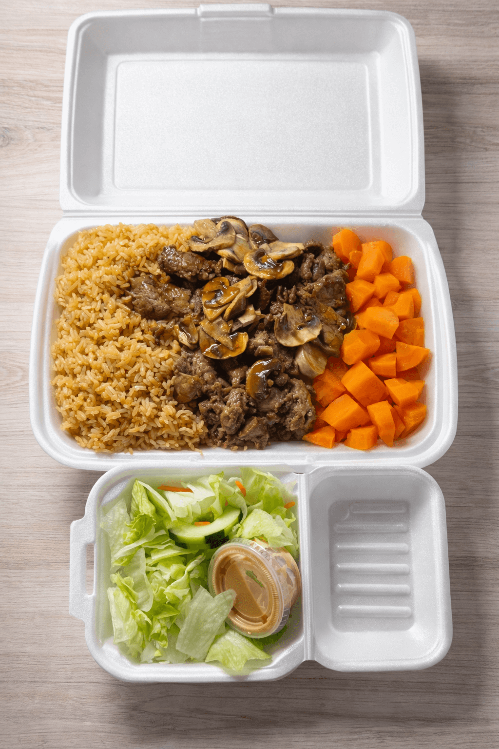 A white styrofoam takeout container with three compartments containing rice, beef with mushrooms, carrots, and a small side salad with dressing.