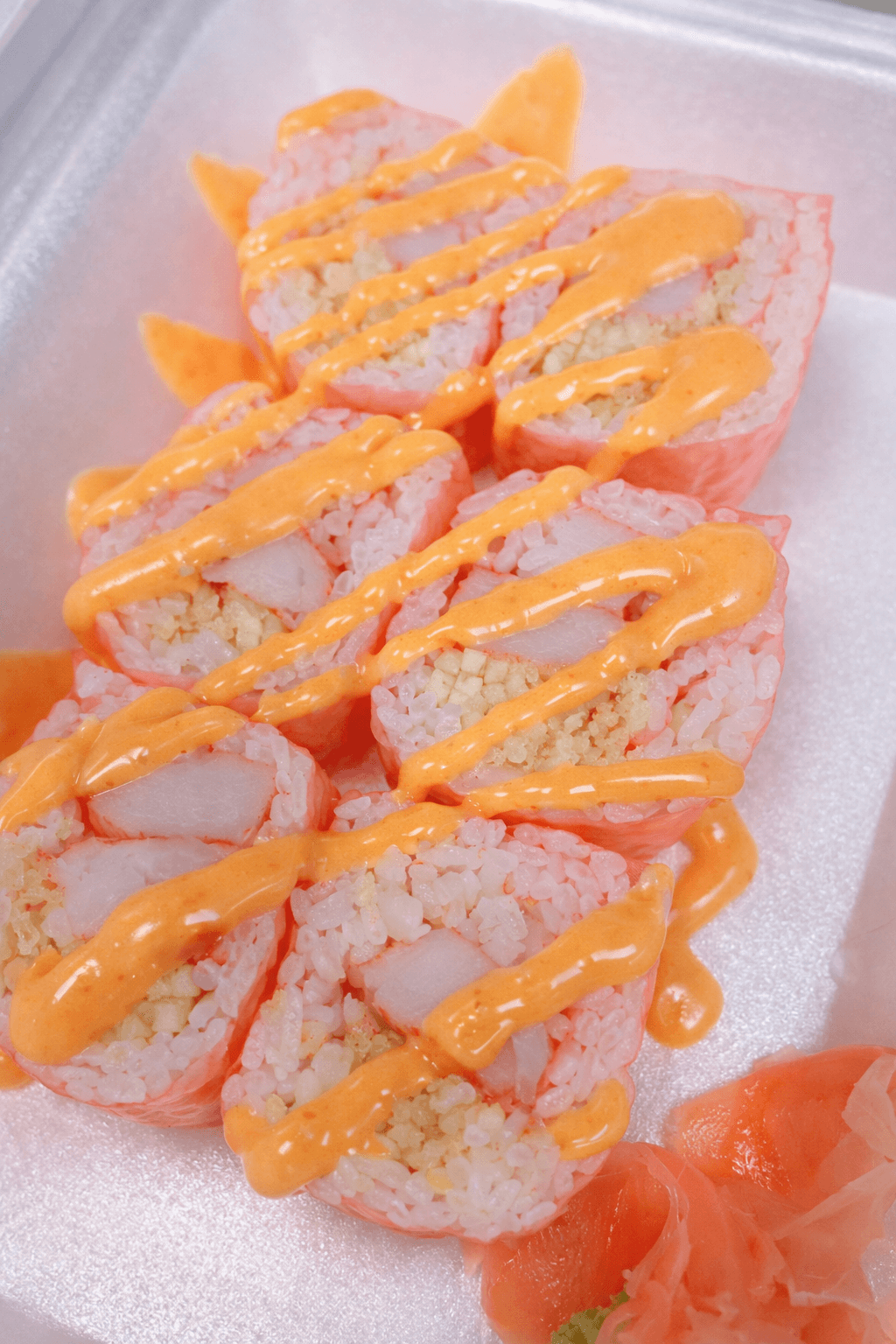 Close-up of sushi rolls drizzled with orange spicy mayo sauce on a white foam tray. There is a side of pickled ginger at the bottom right corner.