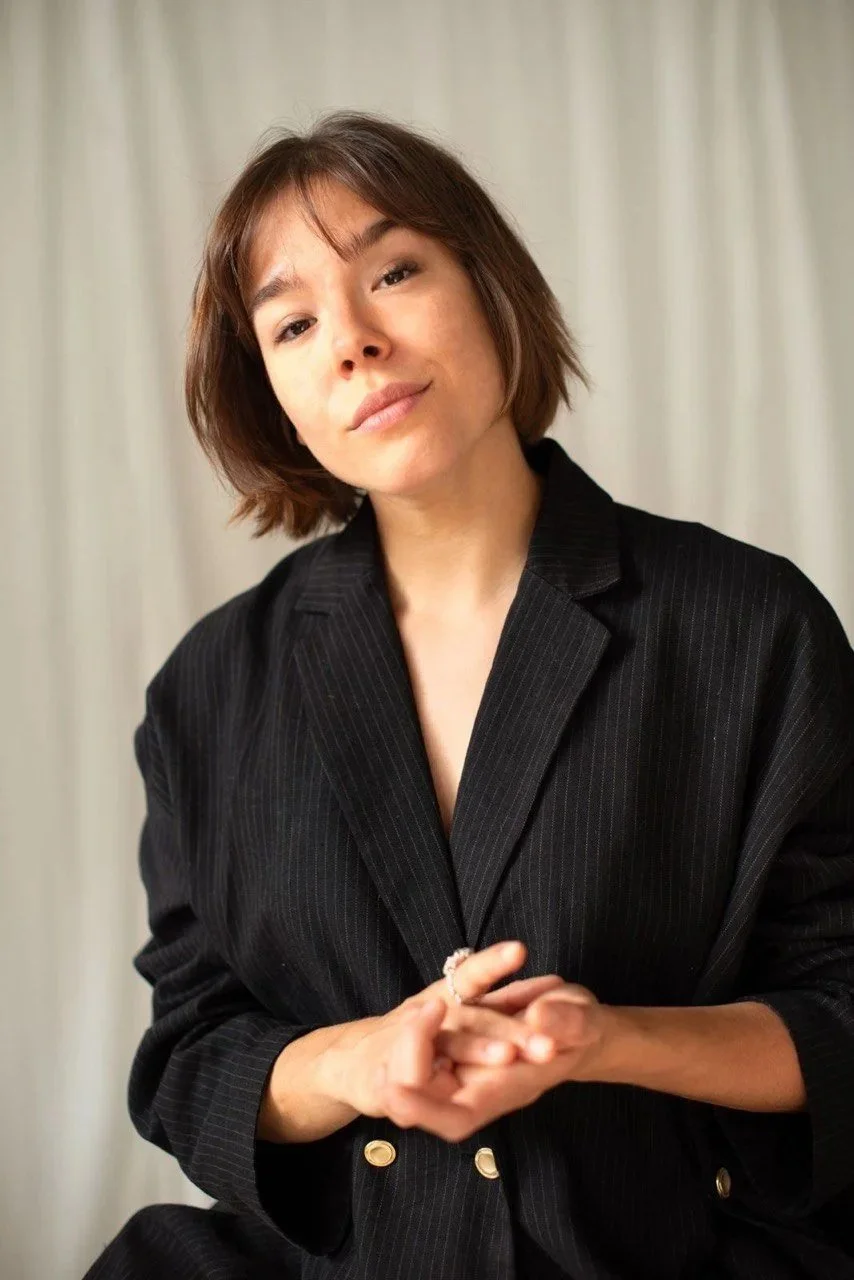 A woman with short brown hair wearing a black pinstriped blazer, sitting in front of white curtains, looking at the camera with a slight smile, clasping her hands.