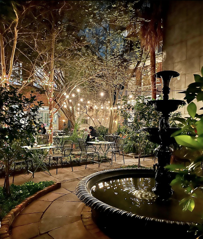 An outdoor restaurant or cafe at night with string lights, trees, and plants, with tables and chairs, and a fountain in the foreground.