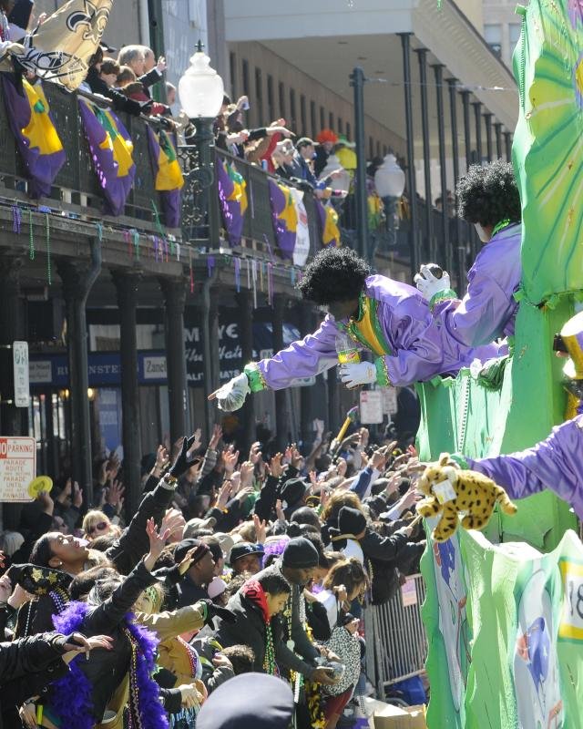 Parade with large float and performers dressed as 1970s disco characters, with a crowd of spectators on balconies and street cheering.