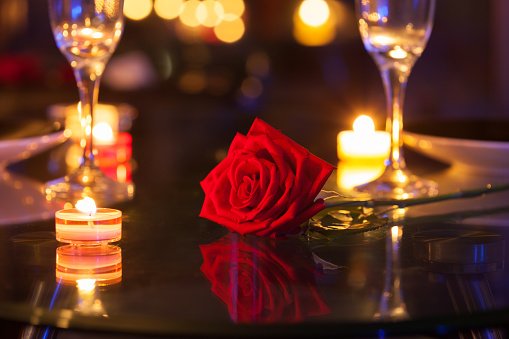 A red rose with green leaves on a dark table, surrounded by lit tea light candles and two empty champagne flutes in a dimly lit setting.