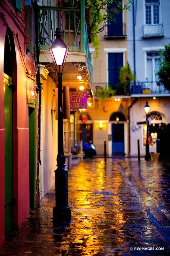 A rainy evening on a colorful European street with wet cobblestones reflecting streetlights, featuring buildings with balconies and a sign indicating an eatery.