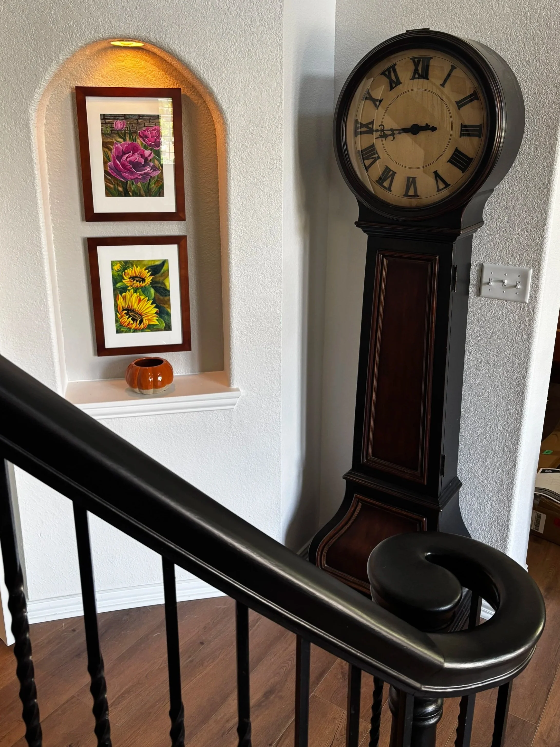 A staircase railing in the foreground with a grandfather clock and art niche in the background, featuring framed floral paintings of sunflowers and pink tulips.