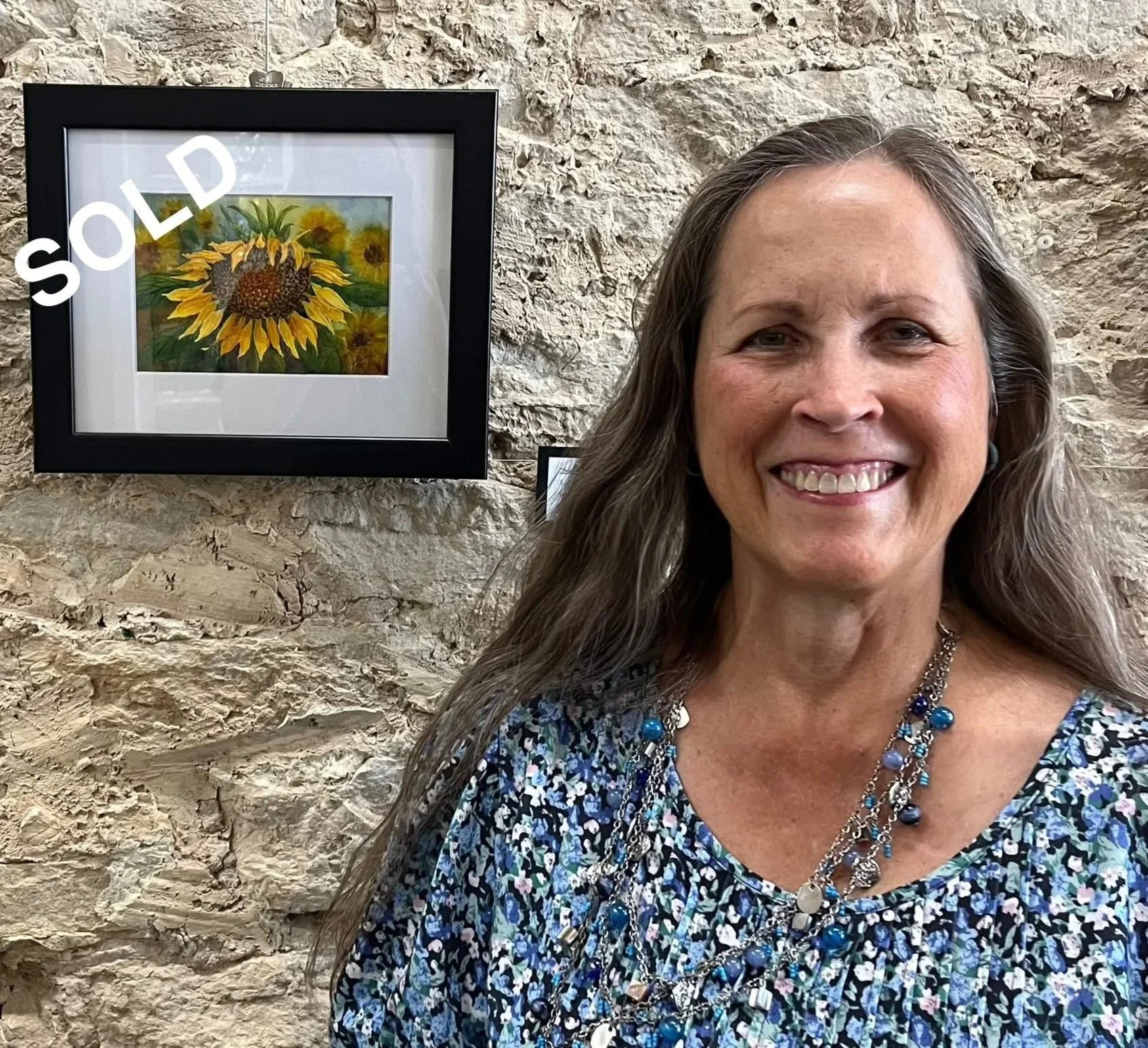 A smiling woman with long gray hair wearing a blue floral shirt and layered necklaces, standing in front of a stone wall with a framed painting of sunflowers.