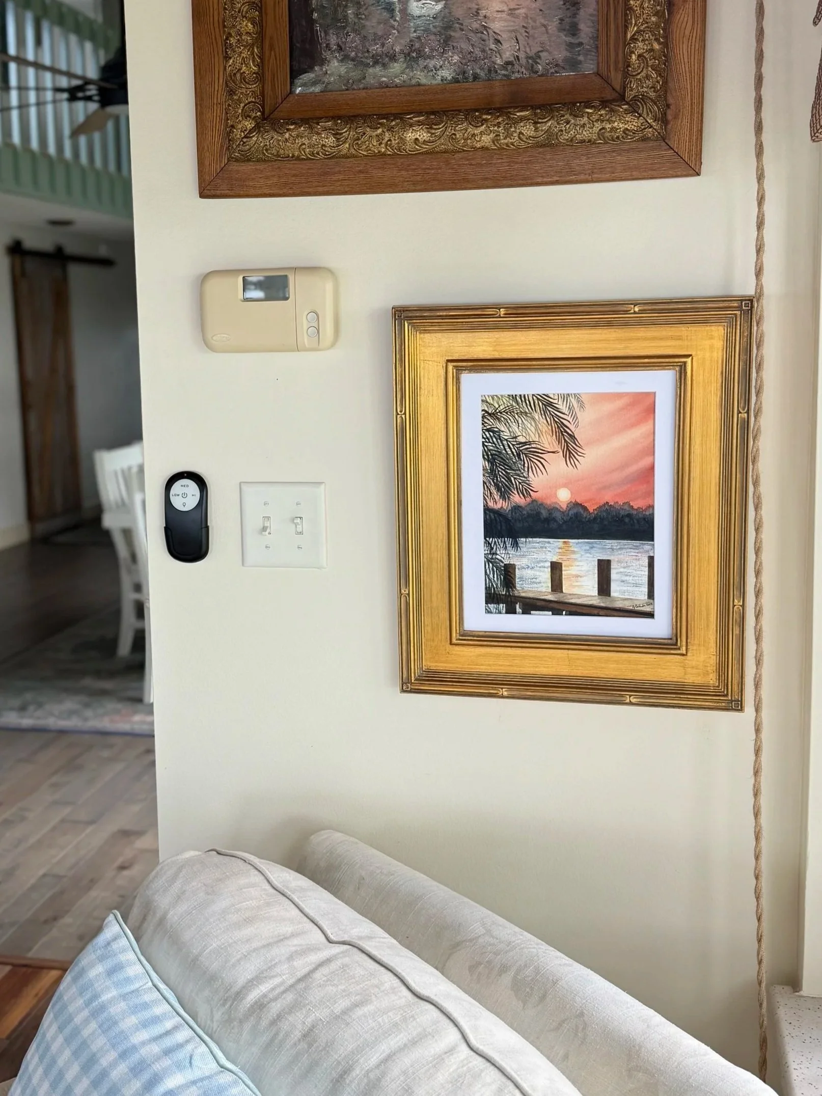 Living room wall with framed landscape painting of a sunset over water, palm trees, and a dock.