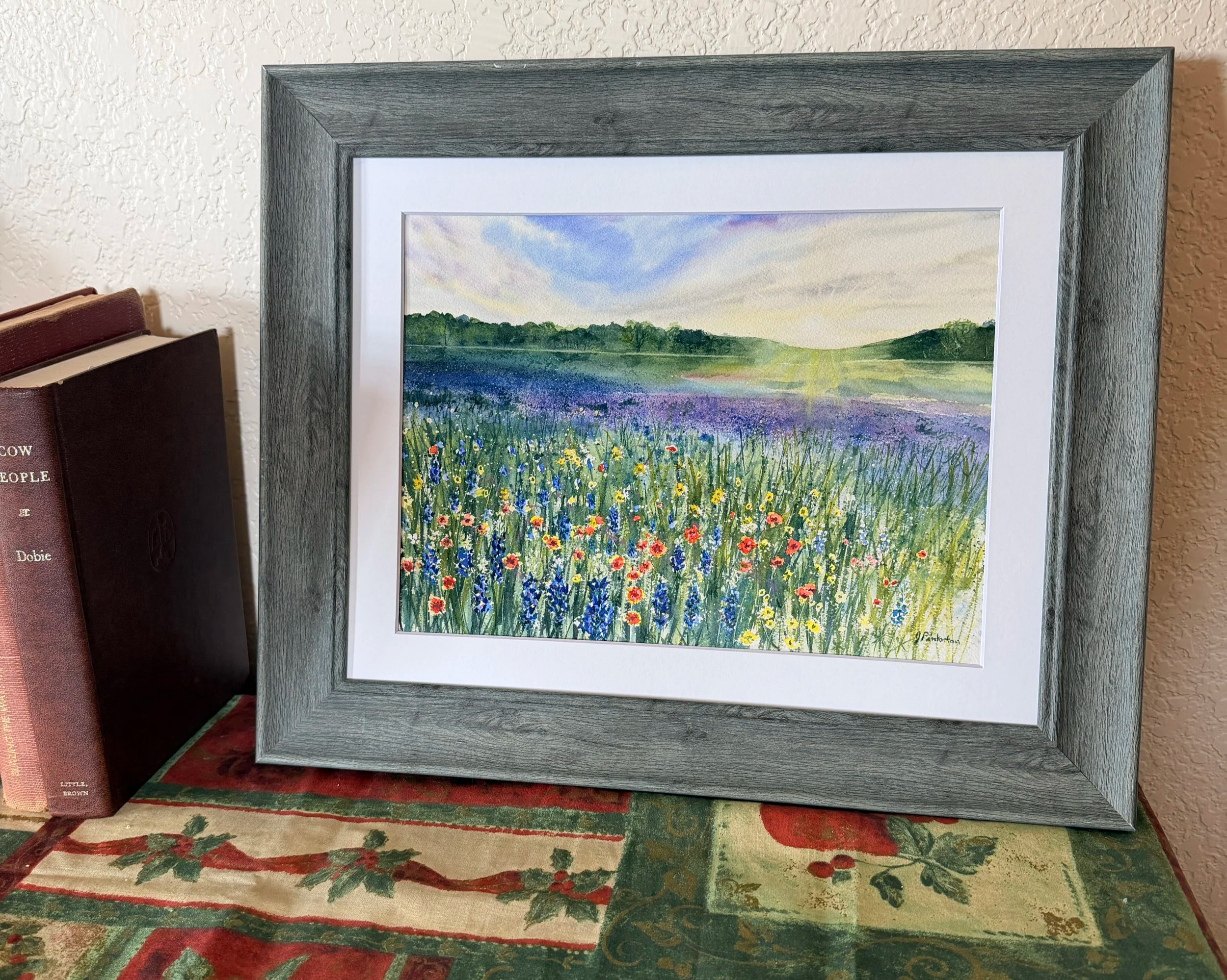A framed watercolor painting of a rising sun lighting up a colorful field of wildflowers with hills in the background, under a partly cloudy sky.