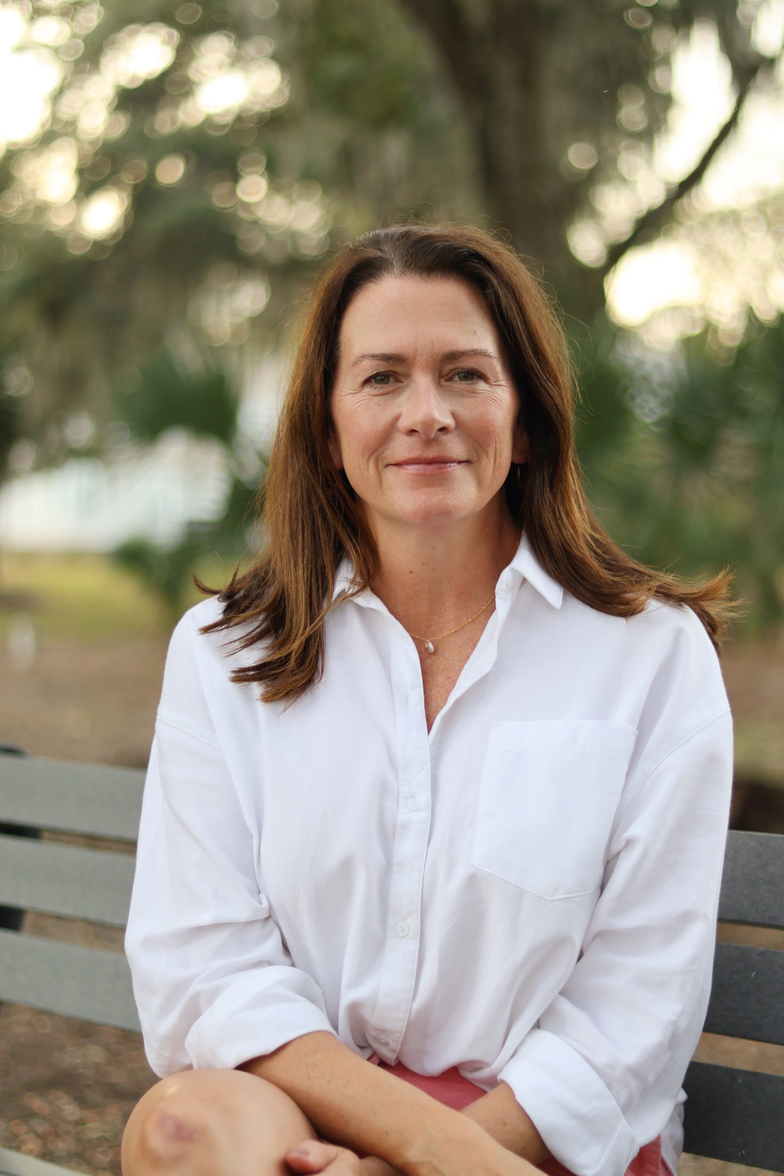 A woman with brown hair sitting on a park bench, wearing a white button-up shirt, with trees and blurred foliage in the background.