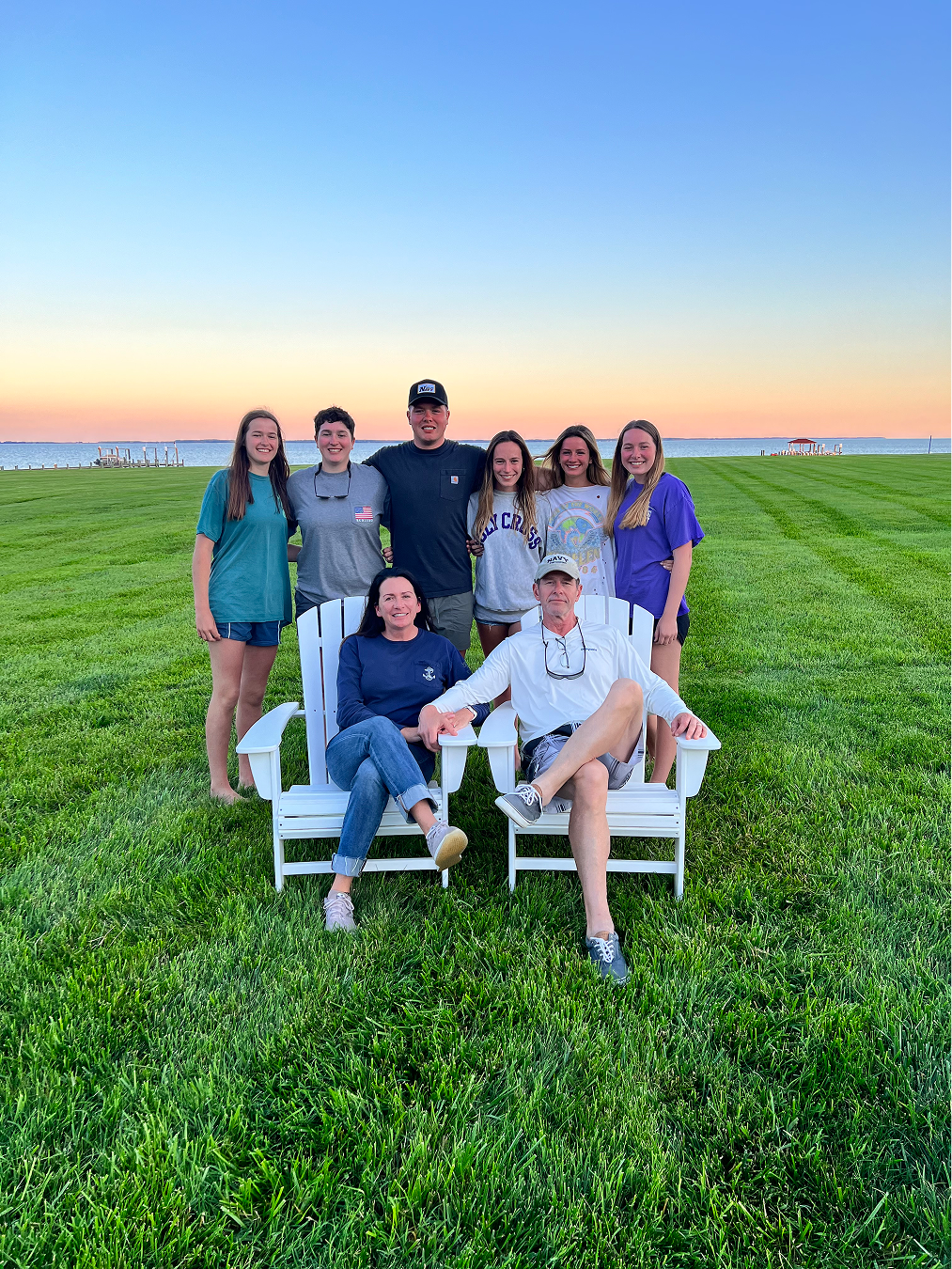 A group of nine people, including teenagers and adults, gathered on a grassy field near a body of water during sunset. Two of the adults are seated on white Adirondack chairs, while the others stand behind them in a line, smiling at the camera.