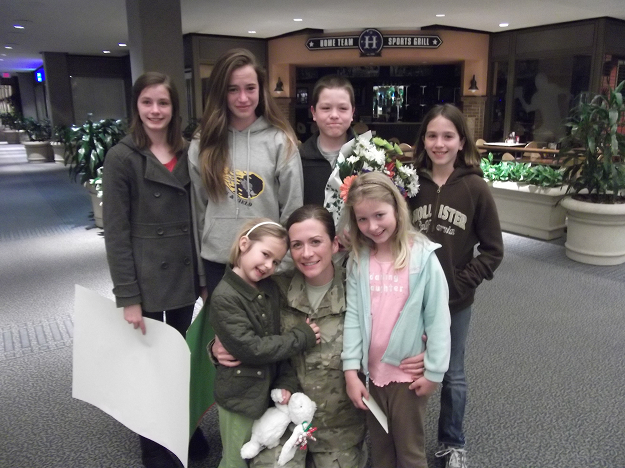 A woman in military uniform posing with six children in a lobby area, some holding flowers and stuffed animals.