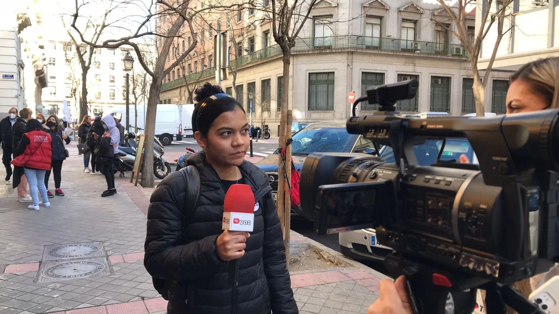 Joven reportera con micrófono en mano siendo grabada por una cámara mientras realiza una noticia en la calle, con varias personas y automóviles al fondo.