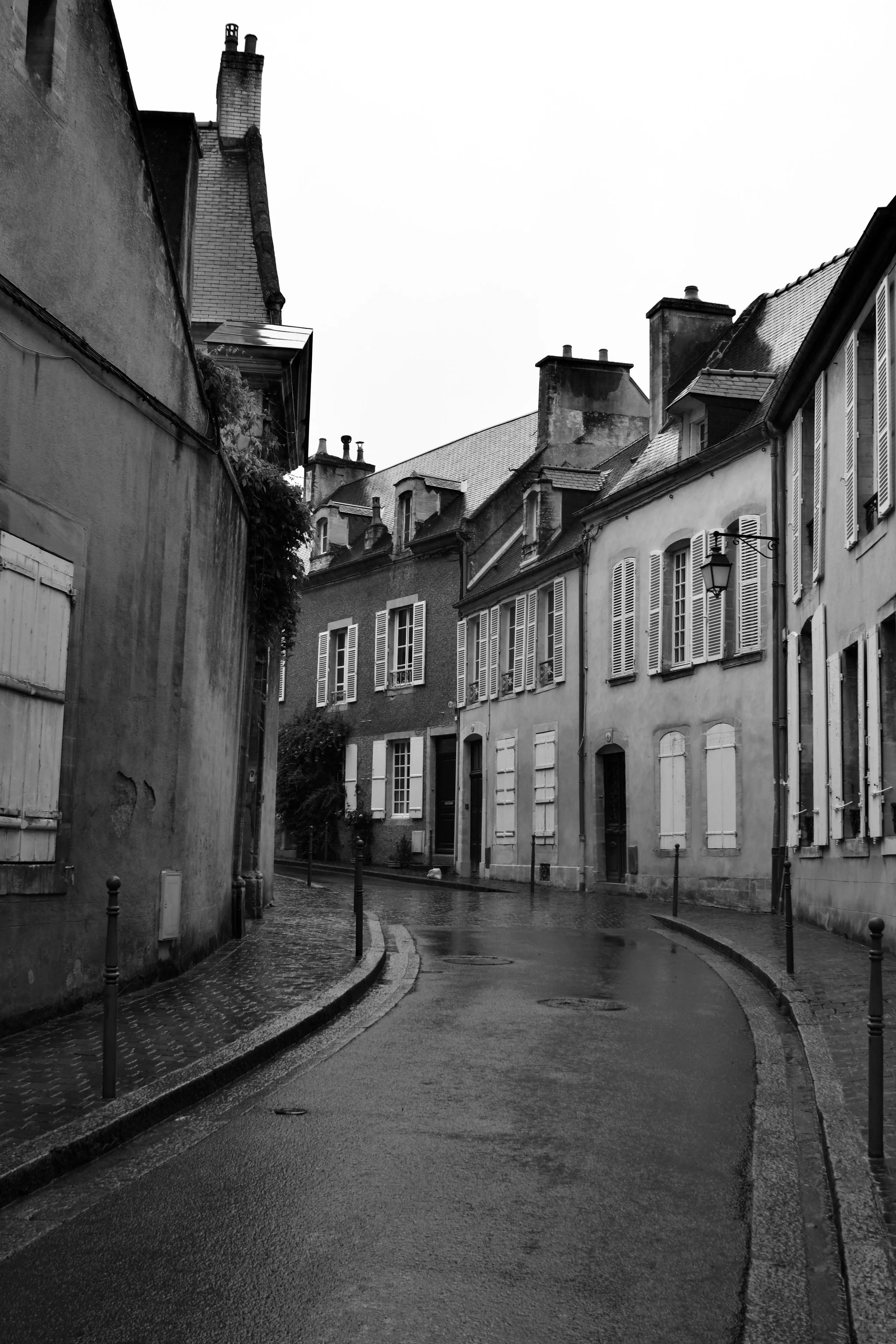 Black and white photo of a narrow, wet, cobblestone street in a European city, lined with old buildings with closed shutters and chimney stacks, on a cloudy day.
