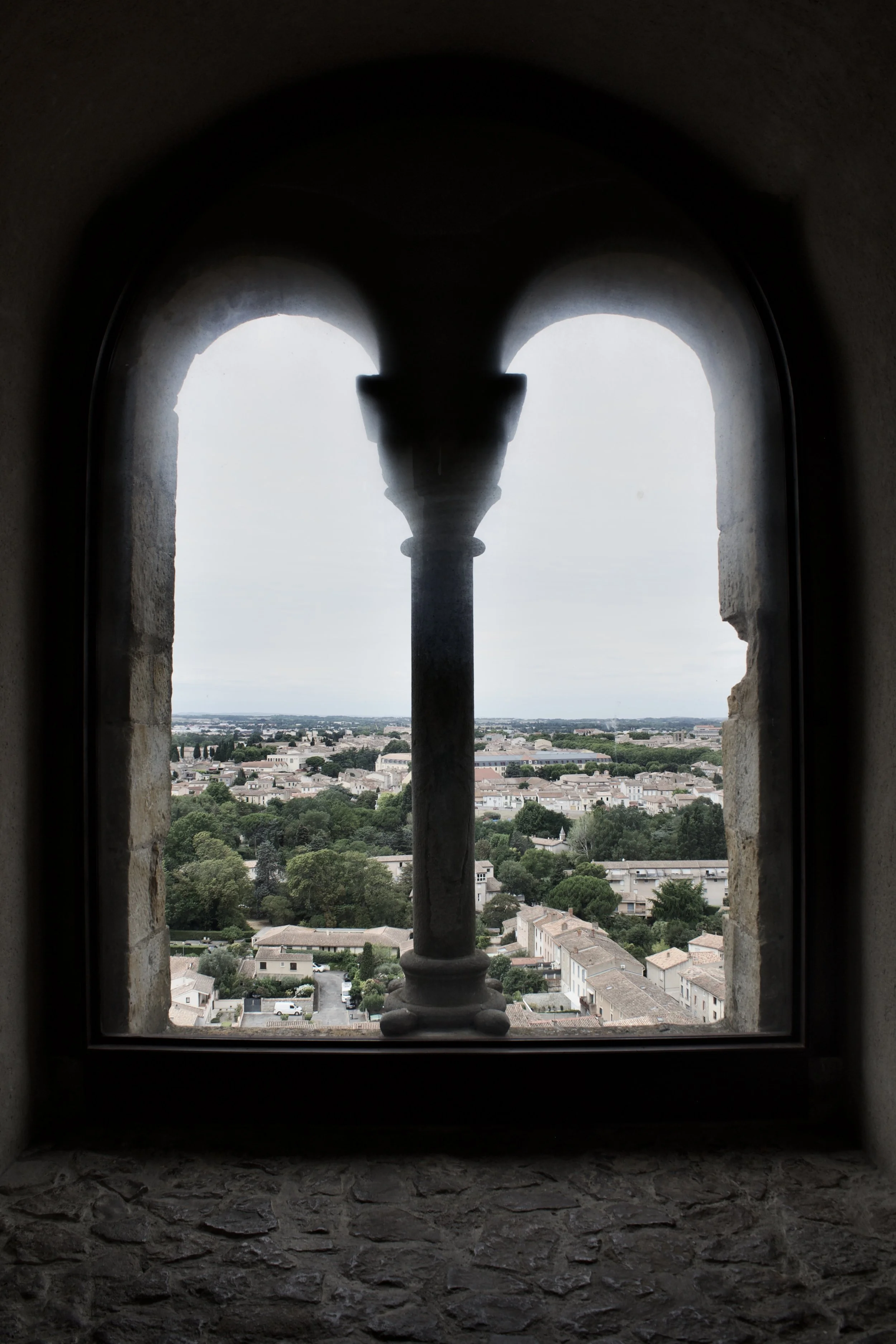 View through a stone-framed window with an ornate column, overlooking a cityscape with buildings, trees, and a cloudy sky.
