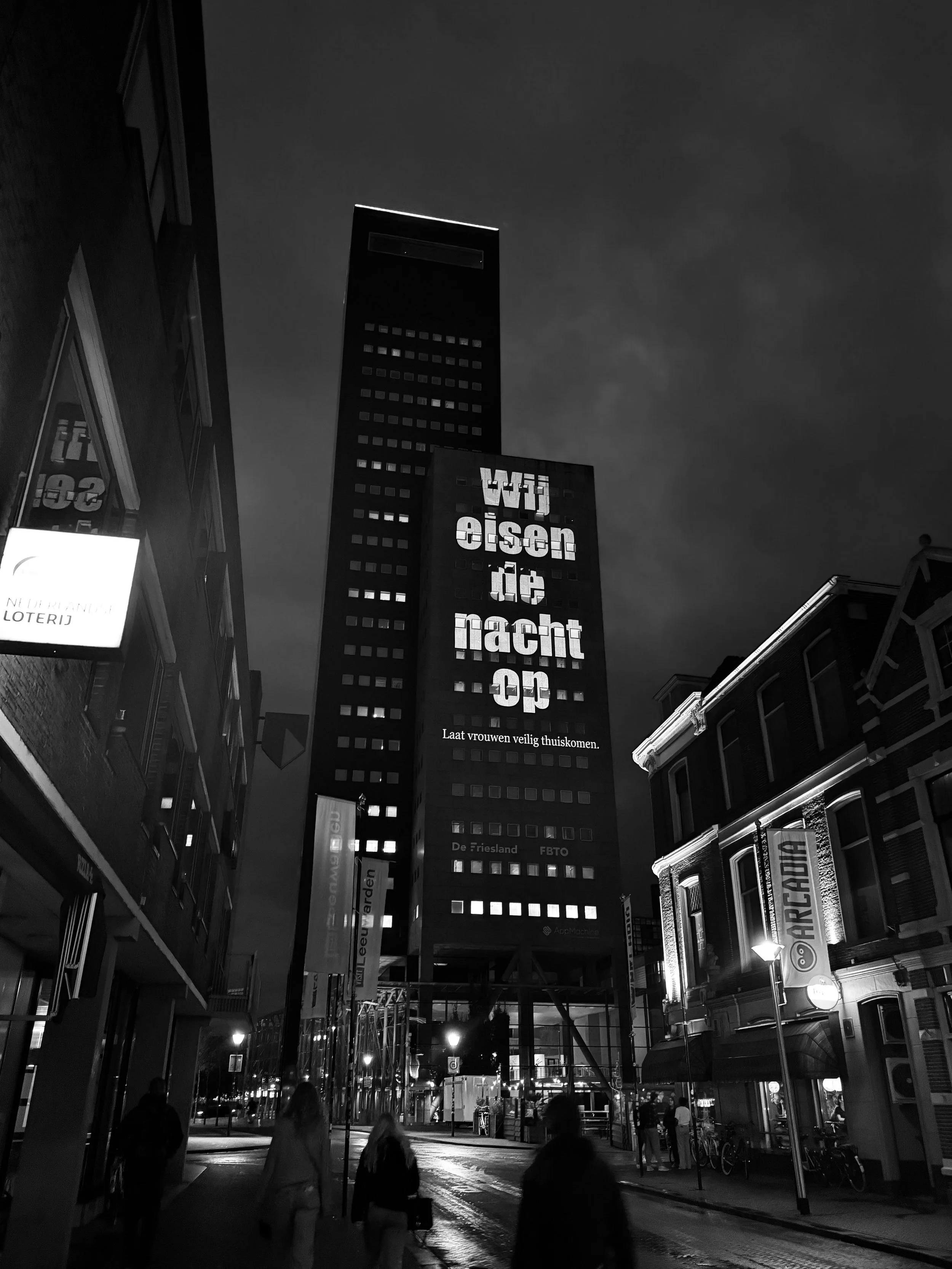 Nighttime city street scene in black and white with tall buildings, pedestrians, and illuminated advertisements on building facades.