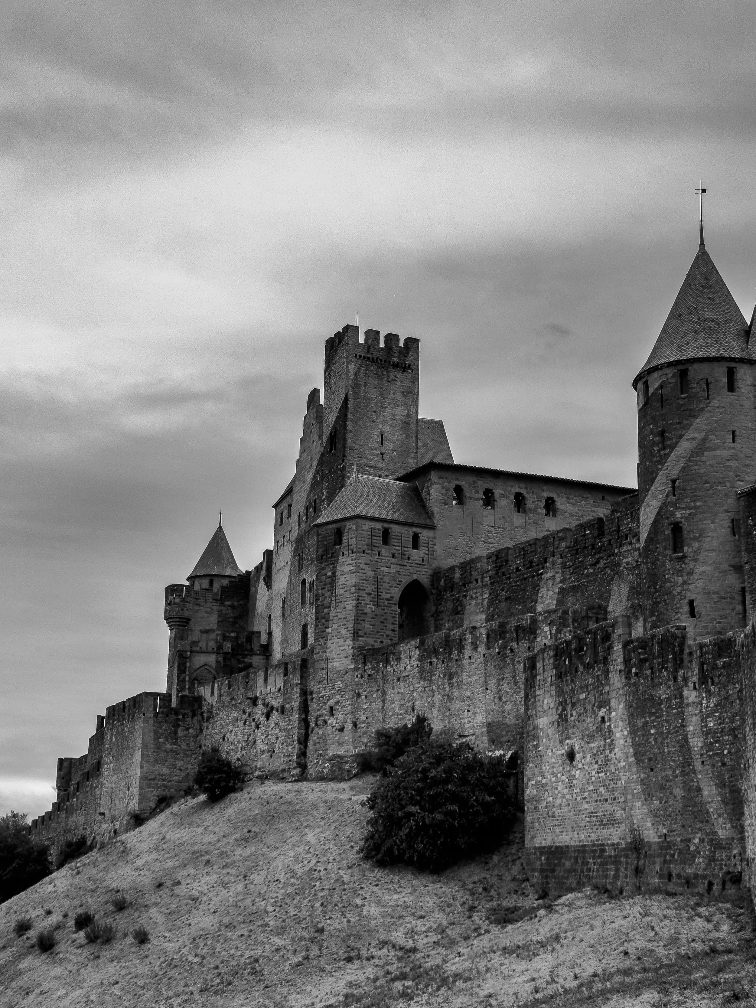 Black and white photograph of a medieval castle with towers, walls, and battlements on a hill, under a cloudy sky.