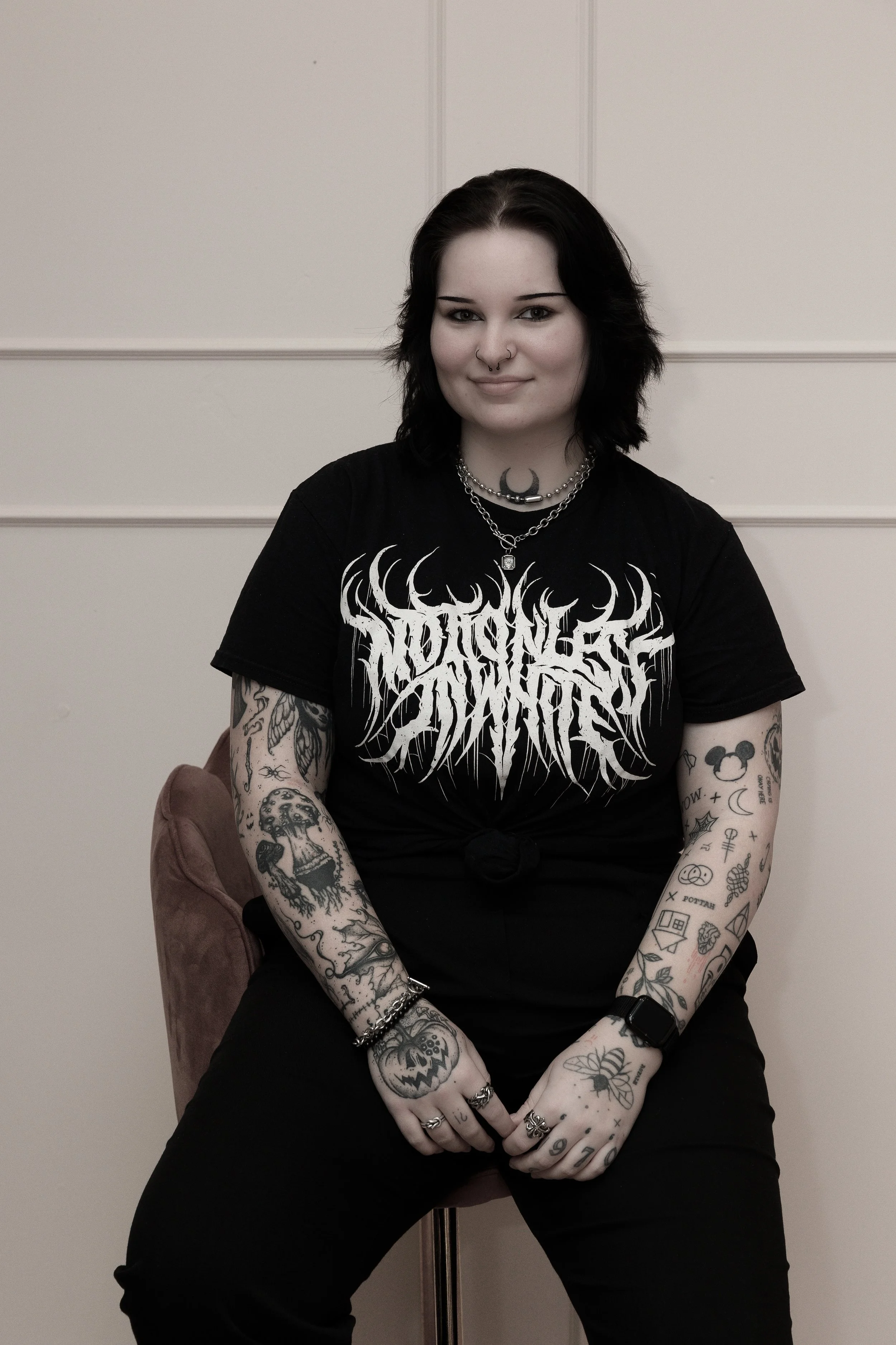 A woman with dark hair, tattoos on her arms, wearing black jewelry and a black graphic T-shirt, sitting on a pink chair in front of a beige wall.