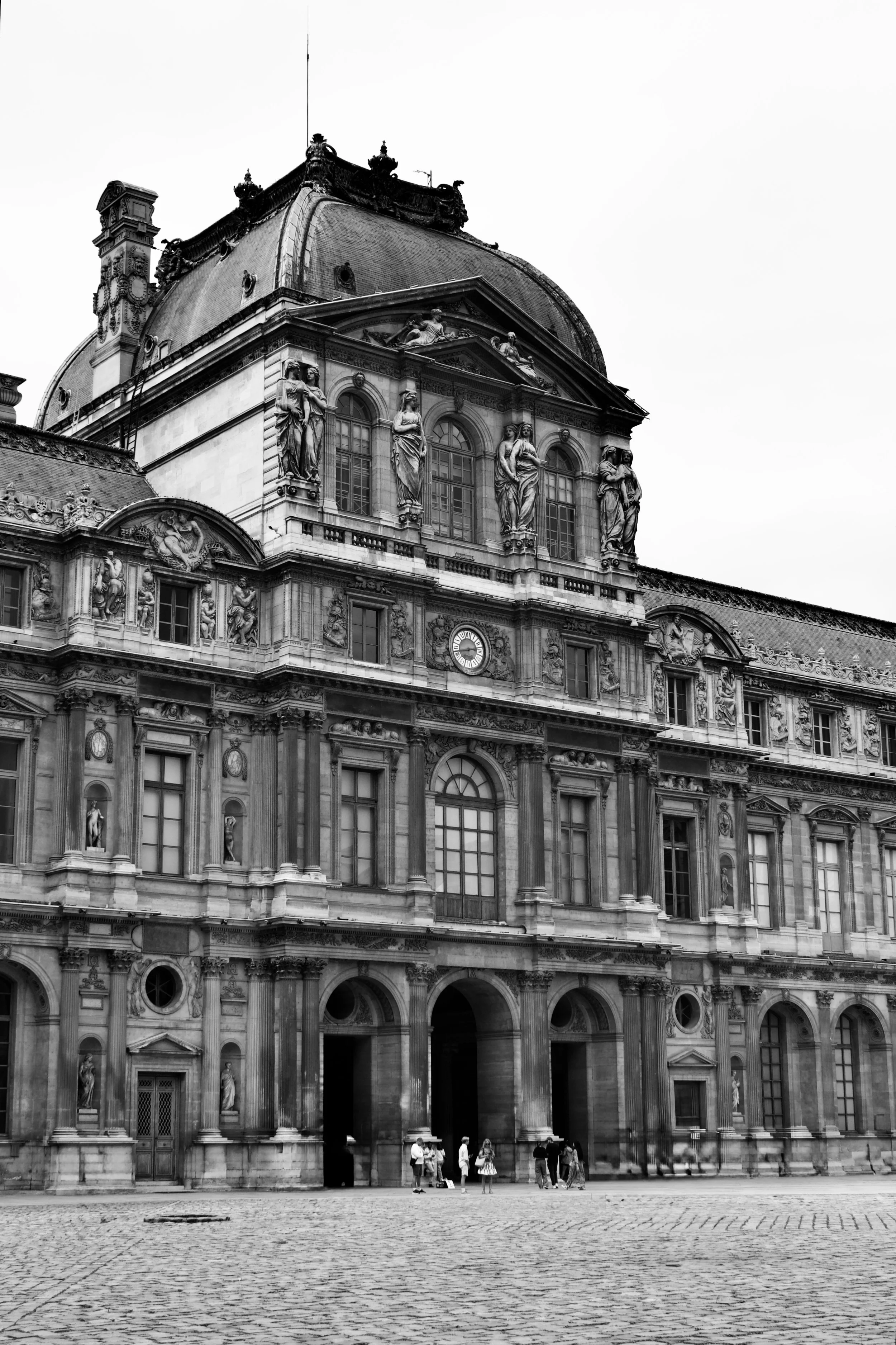 Historical baroque building with ornate sculptures, large windows, and a cobblestone plaza in front, with a group of people standing near the entrance.