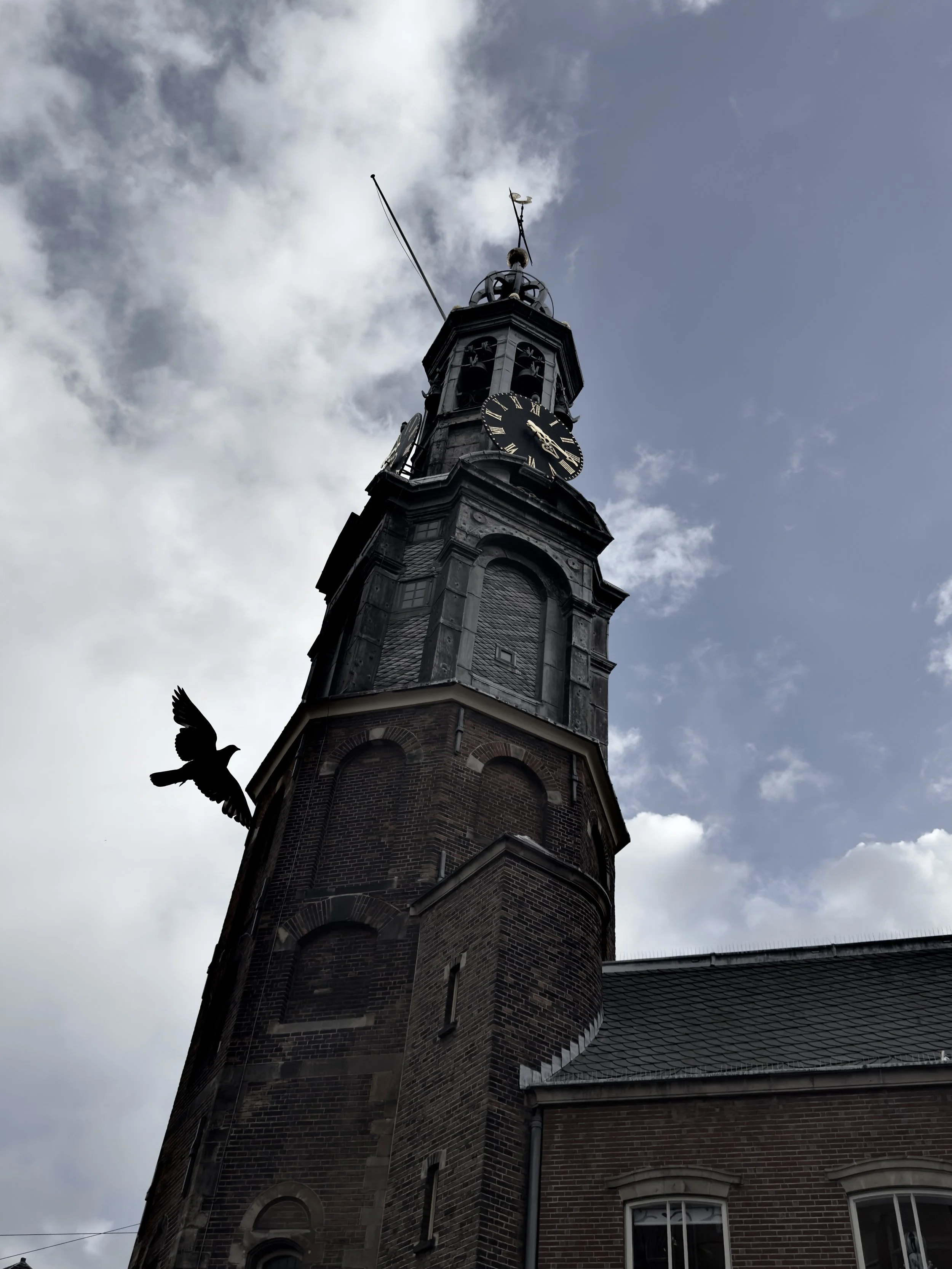 A tall brick church tower with a clock and a weather vane on top, against a partly cloudy sky.