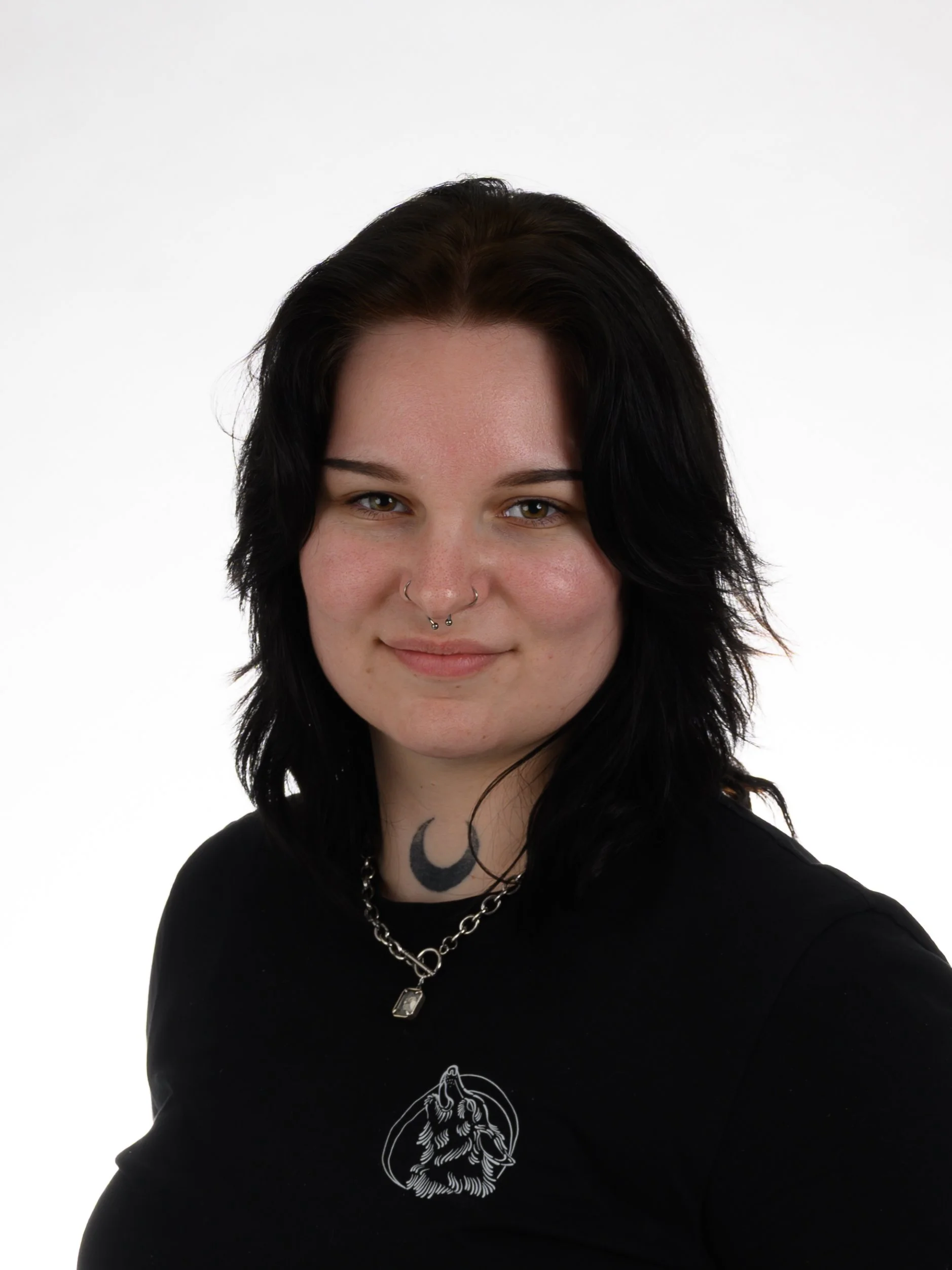 A woman with black, wavy hair, dark eye makeup, multiple facial piercings, a moon tattoo on her neck, and a gothic style necklace. She's wearing a black shirt with a skull graphic and stands against a plain white background.