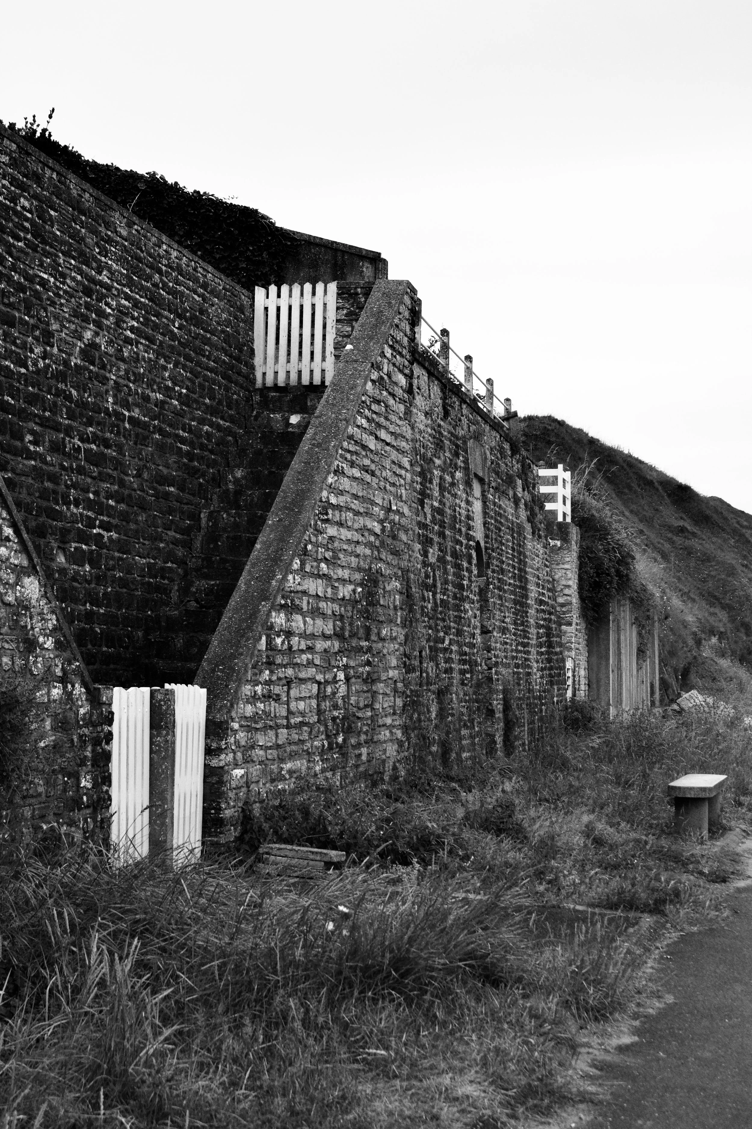 A black and white photo of an old brick sea cliff wall with white fencing on top and a pathway with grass and a bench