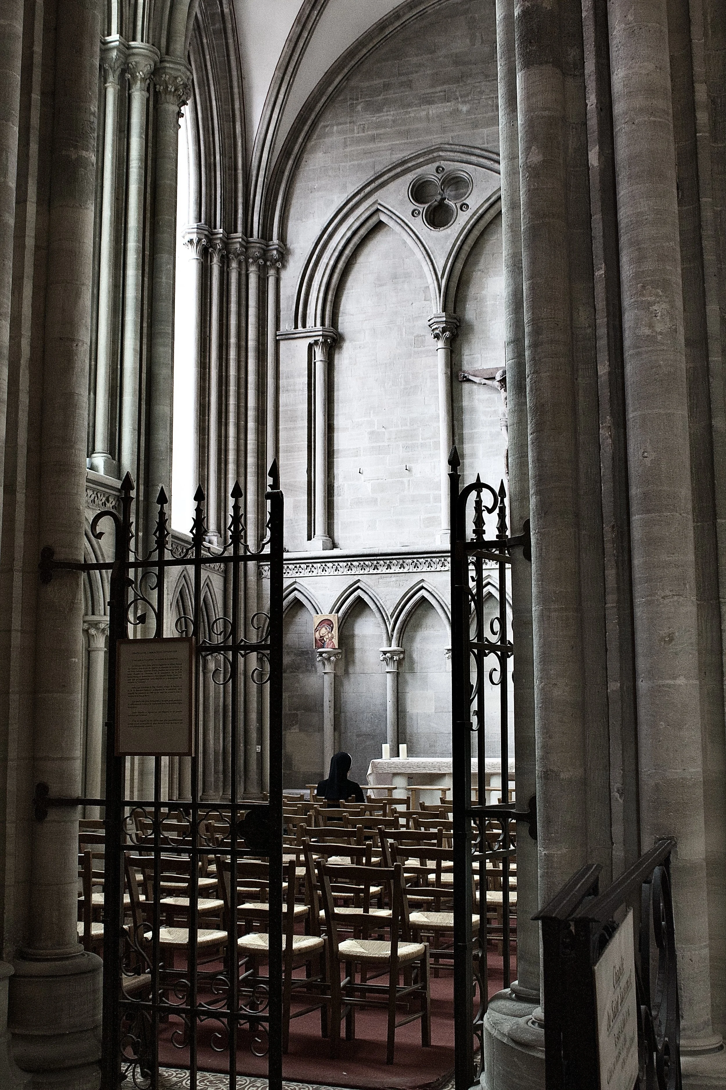 Inside a Catholic church, view through an ornate black wrought iron gate shows wooden chairs arranged in rows, an altar with candles, a painting of Jesus, and a person in black clothing praying, with Gothic-style arches and stone columns in the backg