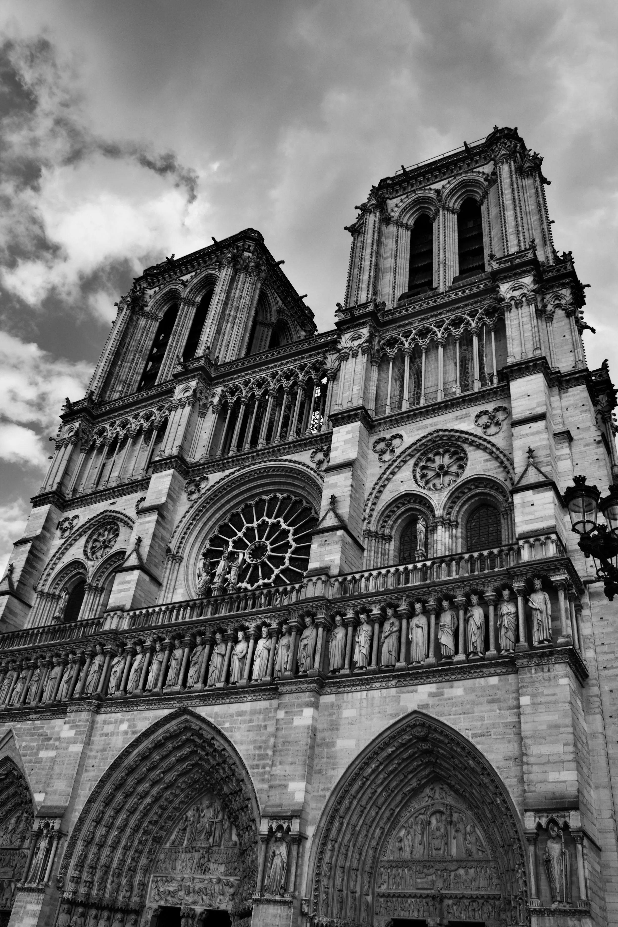 Black and white photo of a Gothic cathedral with two tall bell towers, intricate architectural details, statues, and large arched doorways, with a cloudy sky in the background.