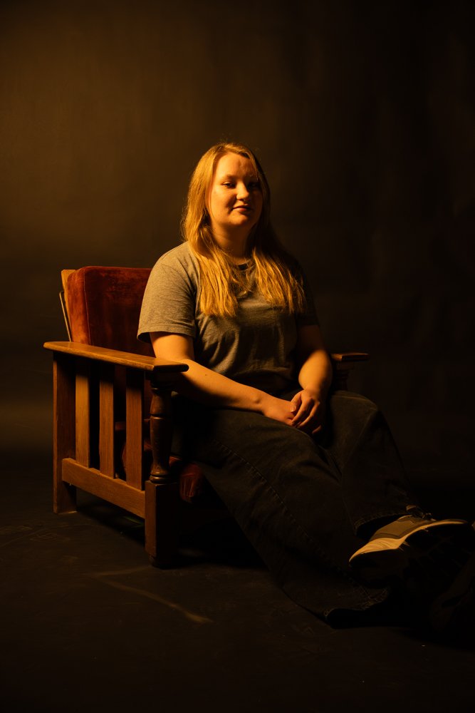 A young woman with long red hair sitting on a wooden chair with a red velvet backrest, on a dark stage with warm lighting.