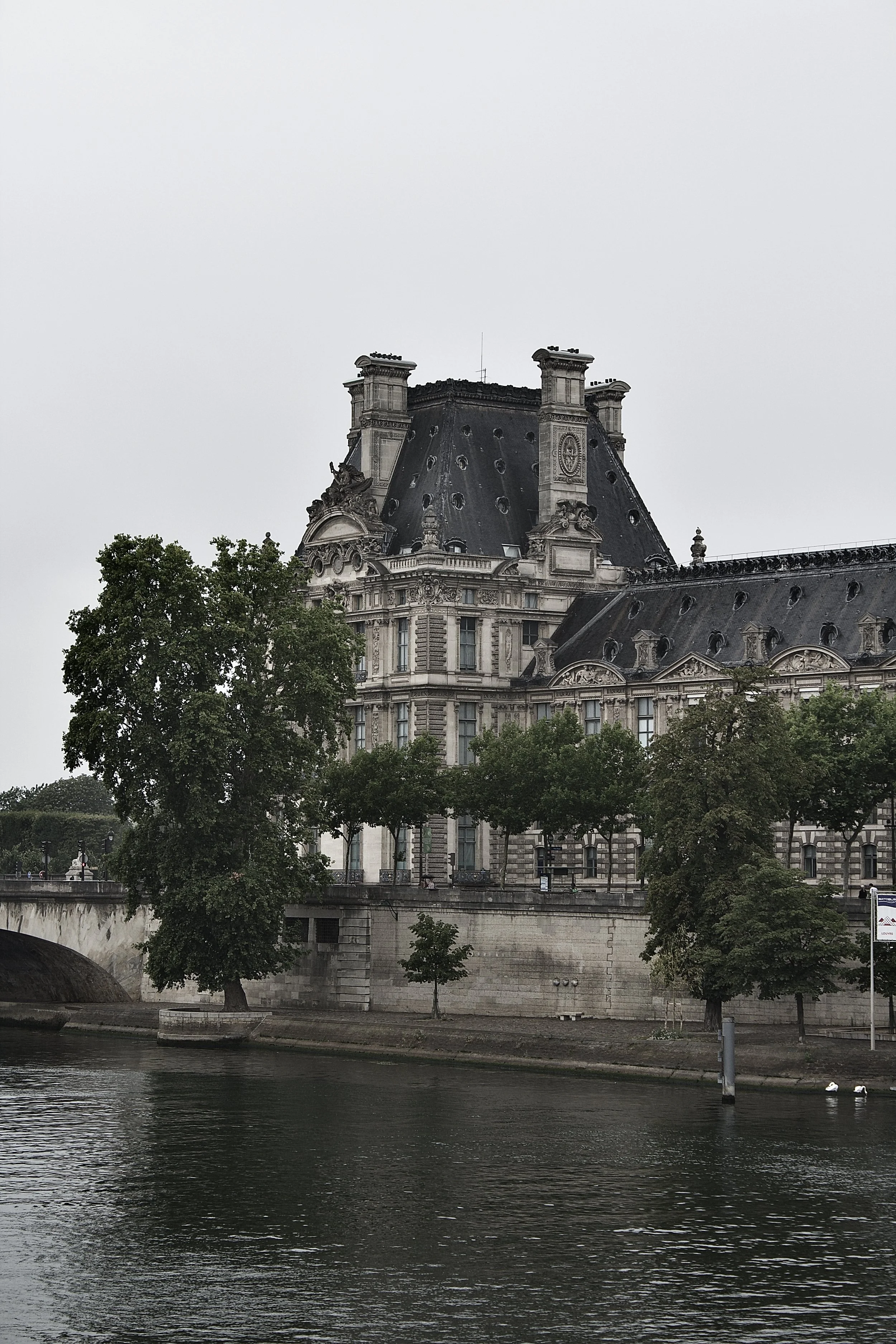 Historic European-style building with ornate architecture, surrounded by green trees and located beside a river under a cloudy sky.