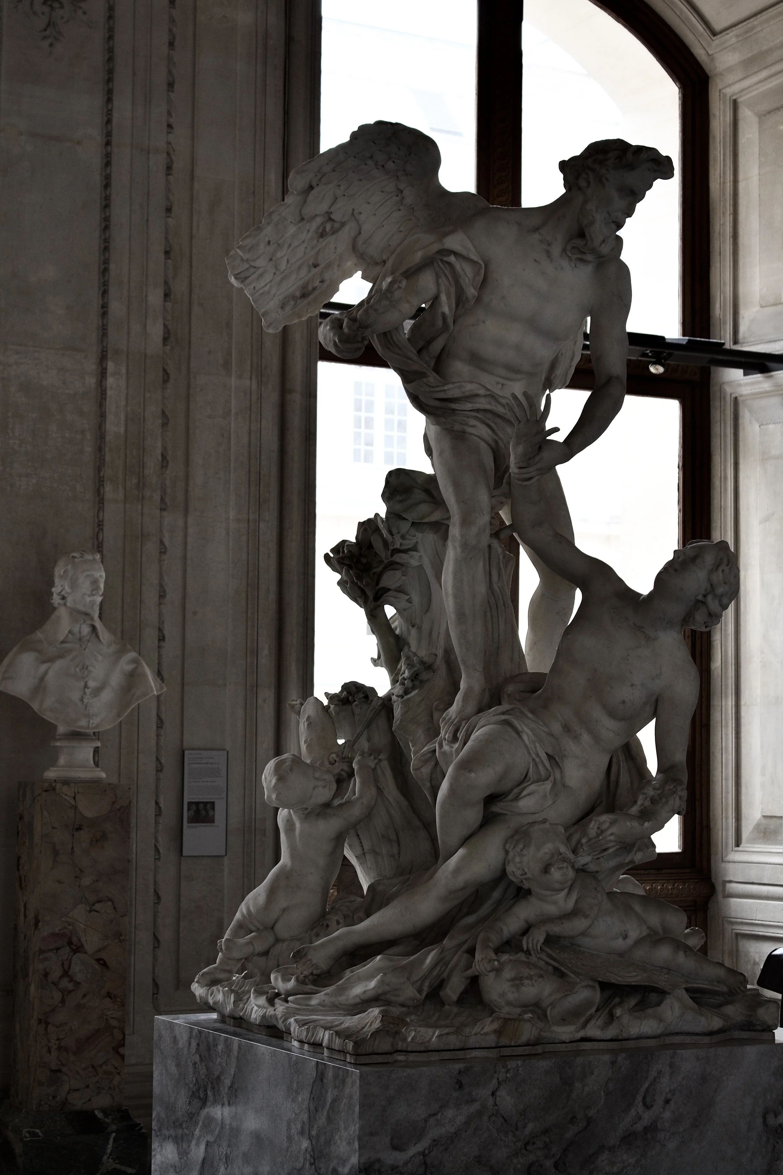 Marble sculpture depicting a mythological scene with a muscular man with wings holding another man, set inside a room with a marble bust on a pedestal and a window in the background.
