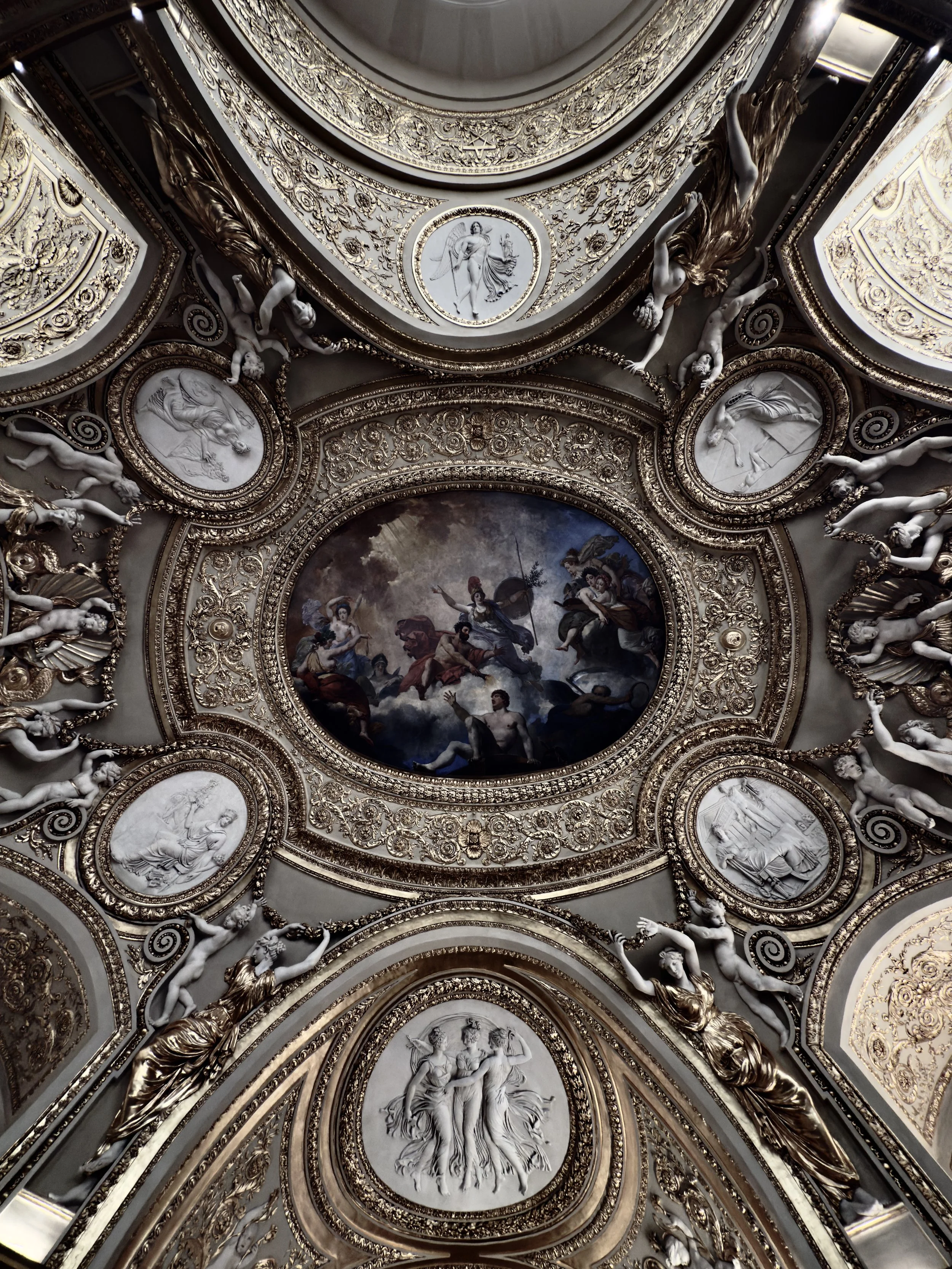 Intricate ceiling with gold ornate moldings, sculptures of reclining figures, and framed bas-relief sculptures surrounding a central painted oval depicting a classical mythological scene with gods and seated figures.