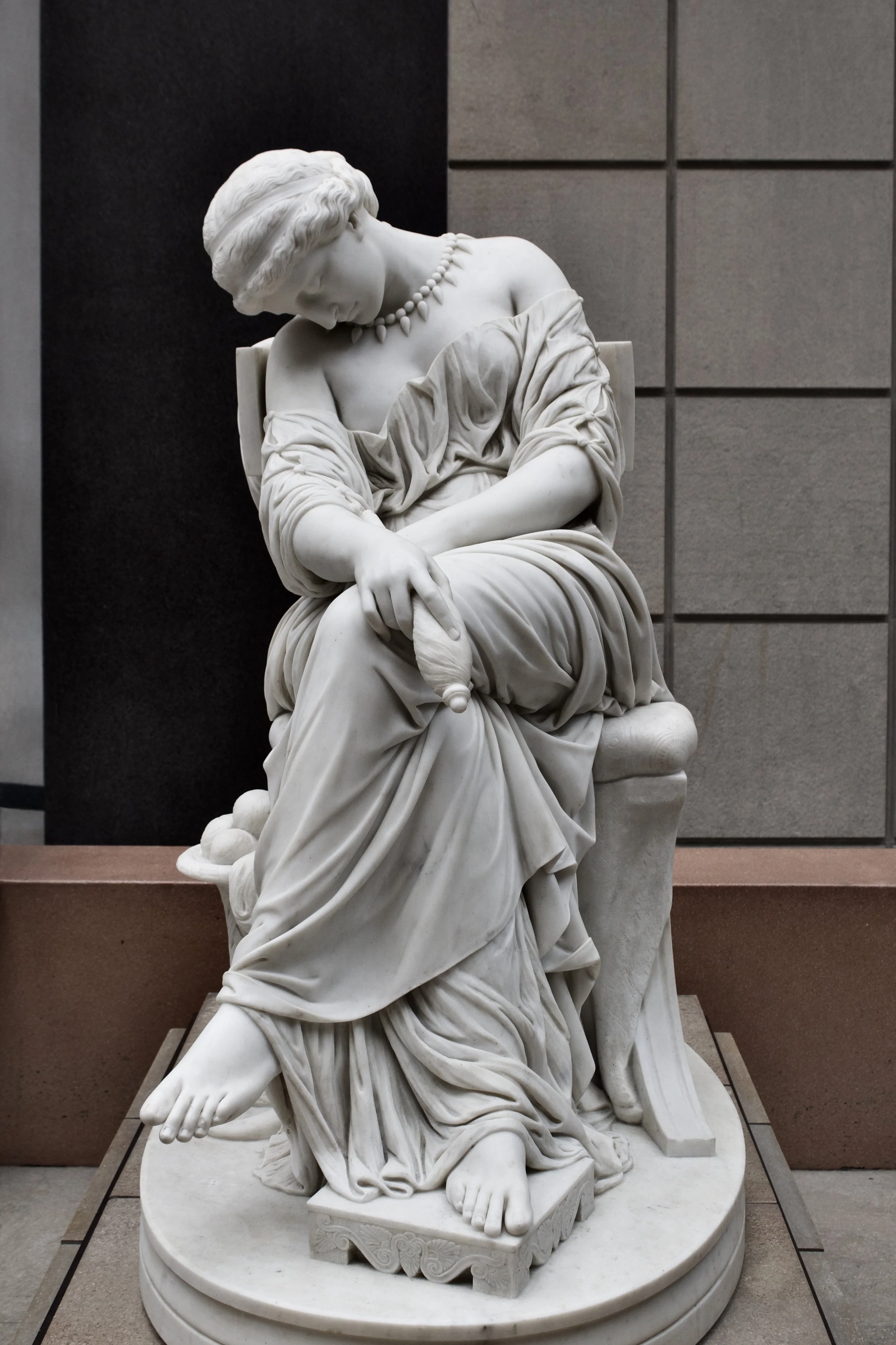 Marble sculpture of a seated woman with head bowed, wearing a draped dress and a necklace, holding a small object in her right hand, displayed indoors against a background of tiled walls.