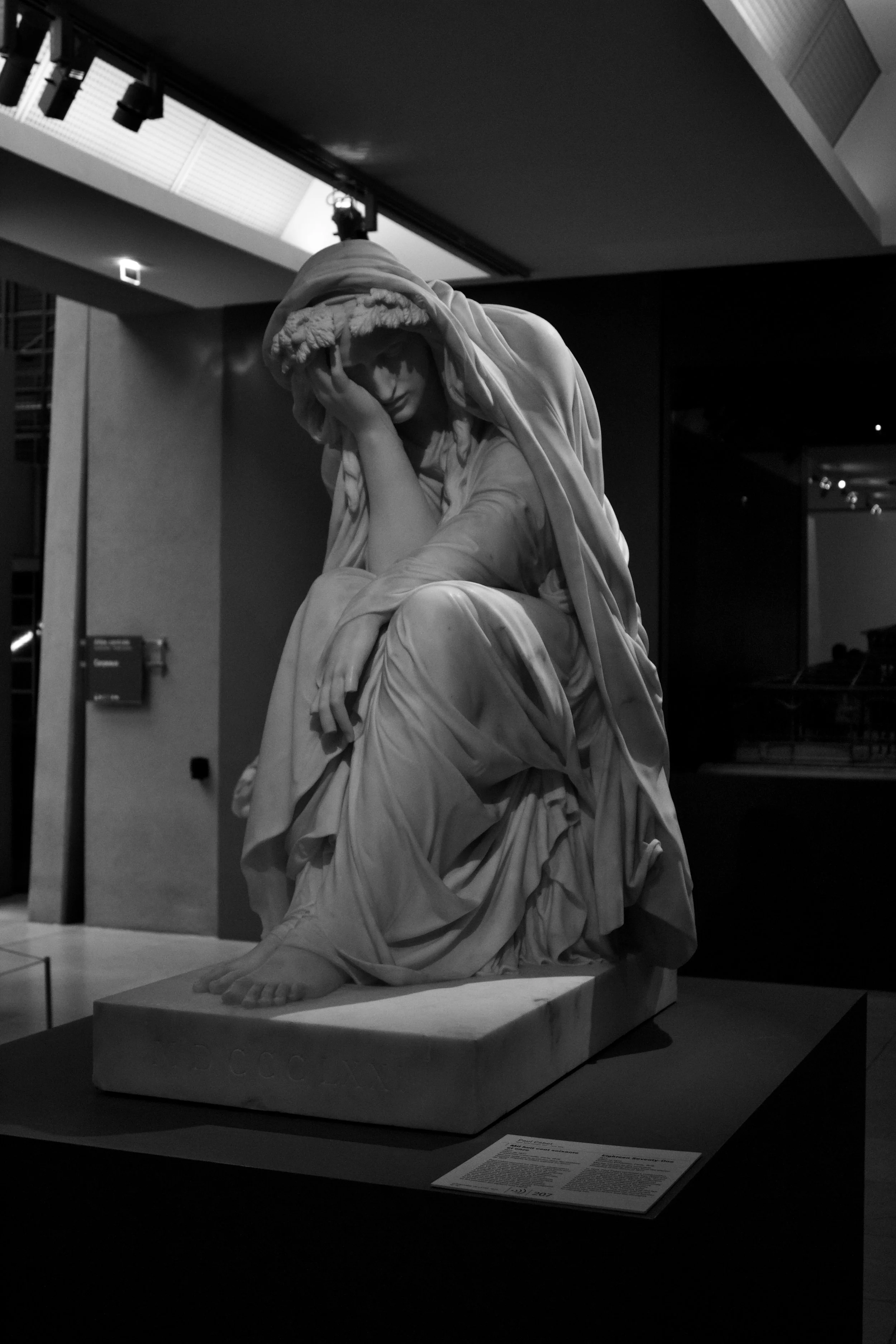 Black and white photograph of a classical marble sculpture depicting a seated woman with her head resting on her hand, wearing a hooded cloak, displayed in a museum.