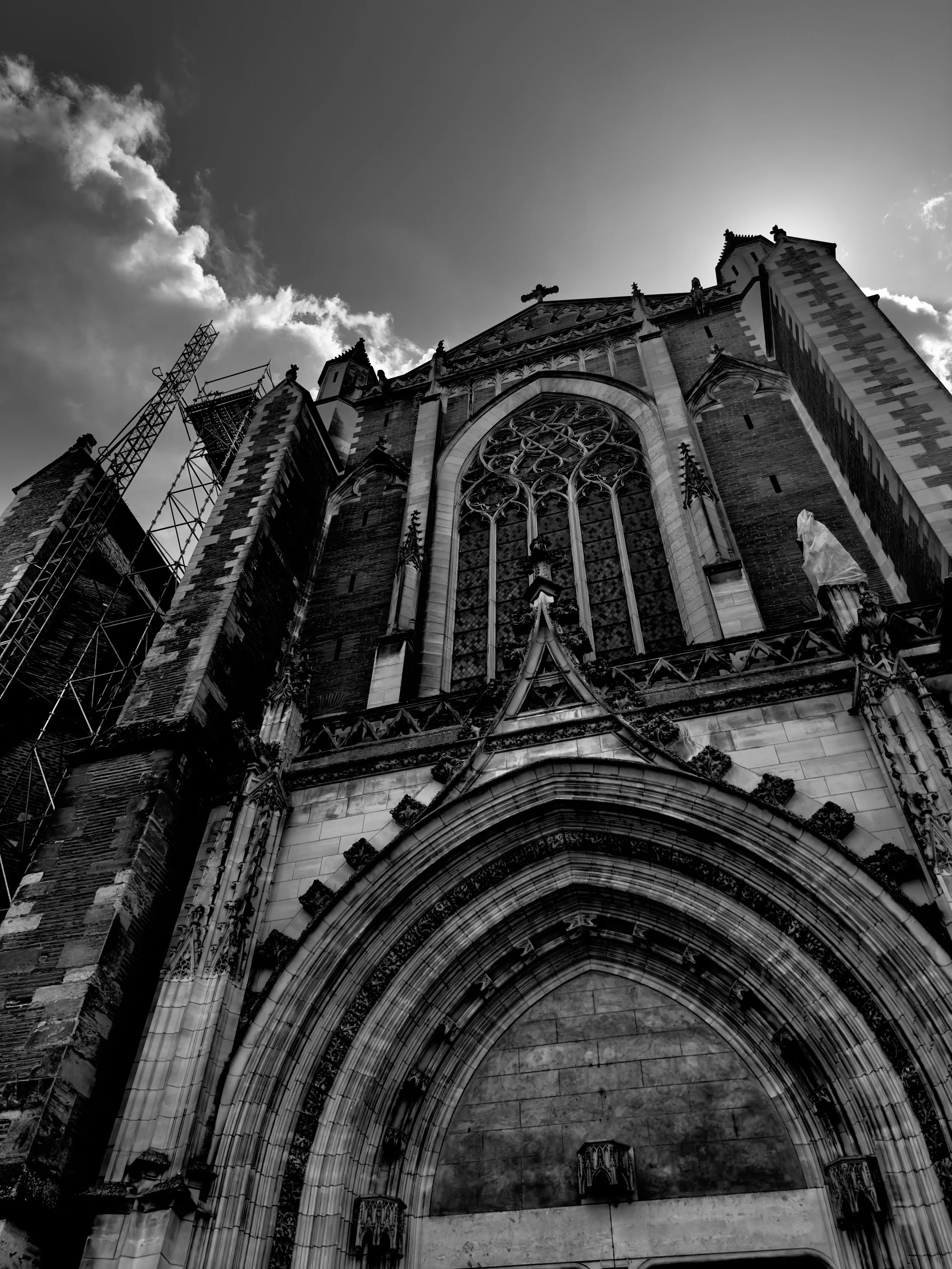 Black and white photo of a Gothic cathedral viewed from below, showing detailed stone architecture, large stained glass window, and a cloudy sky in the background.