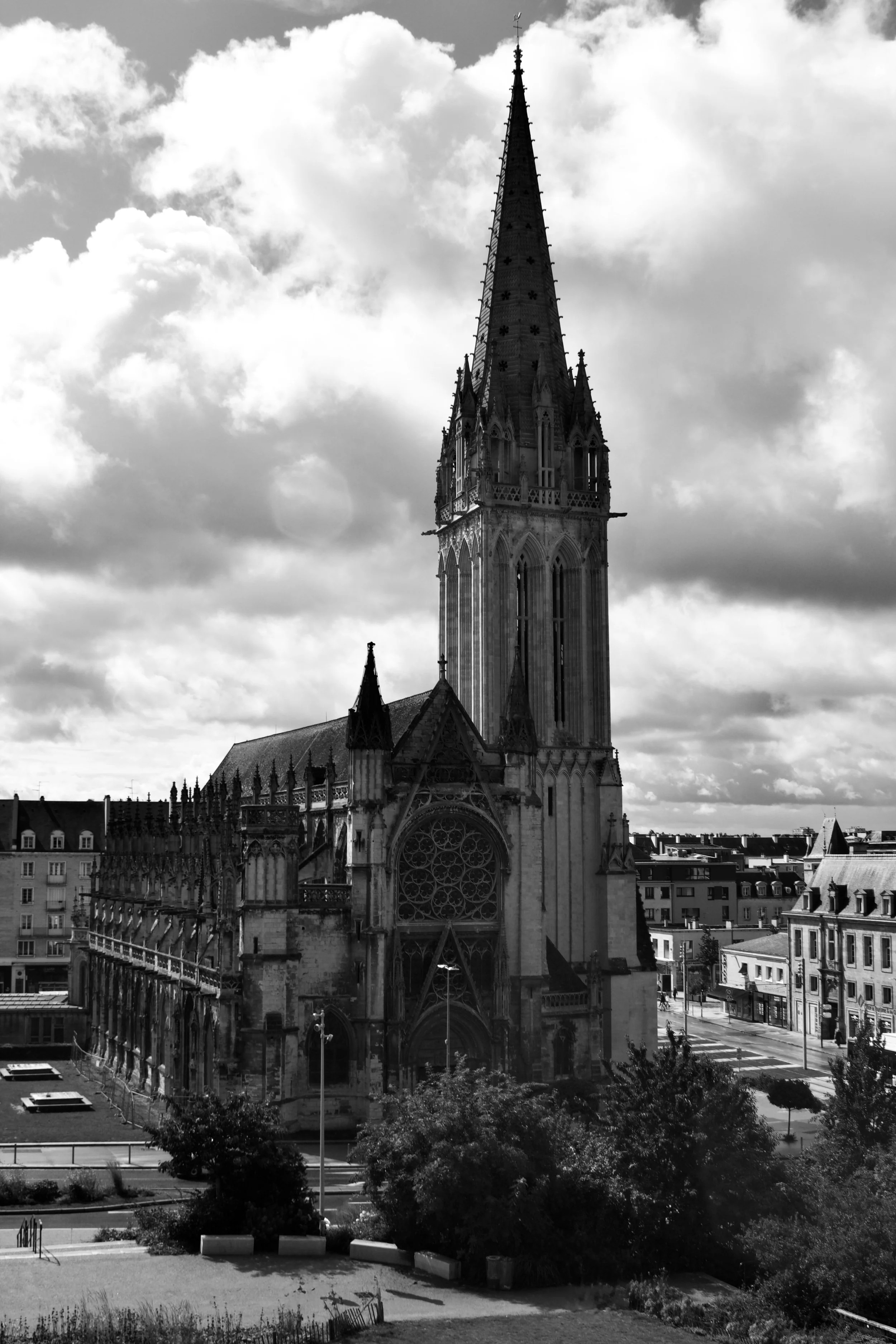 Black and white photo of a tall Gothic-style church with a prominent spire, set against a partly cloudy sky, with surrounding city buildings and trees.