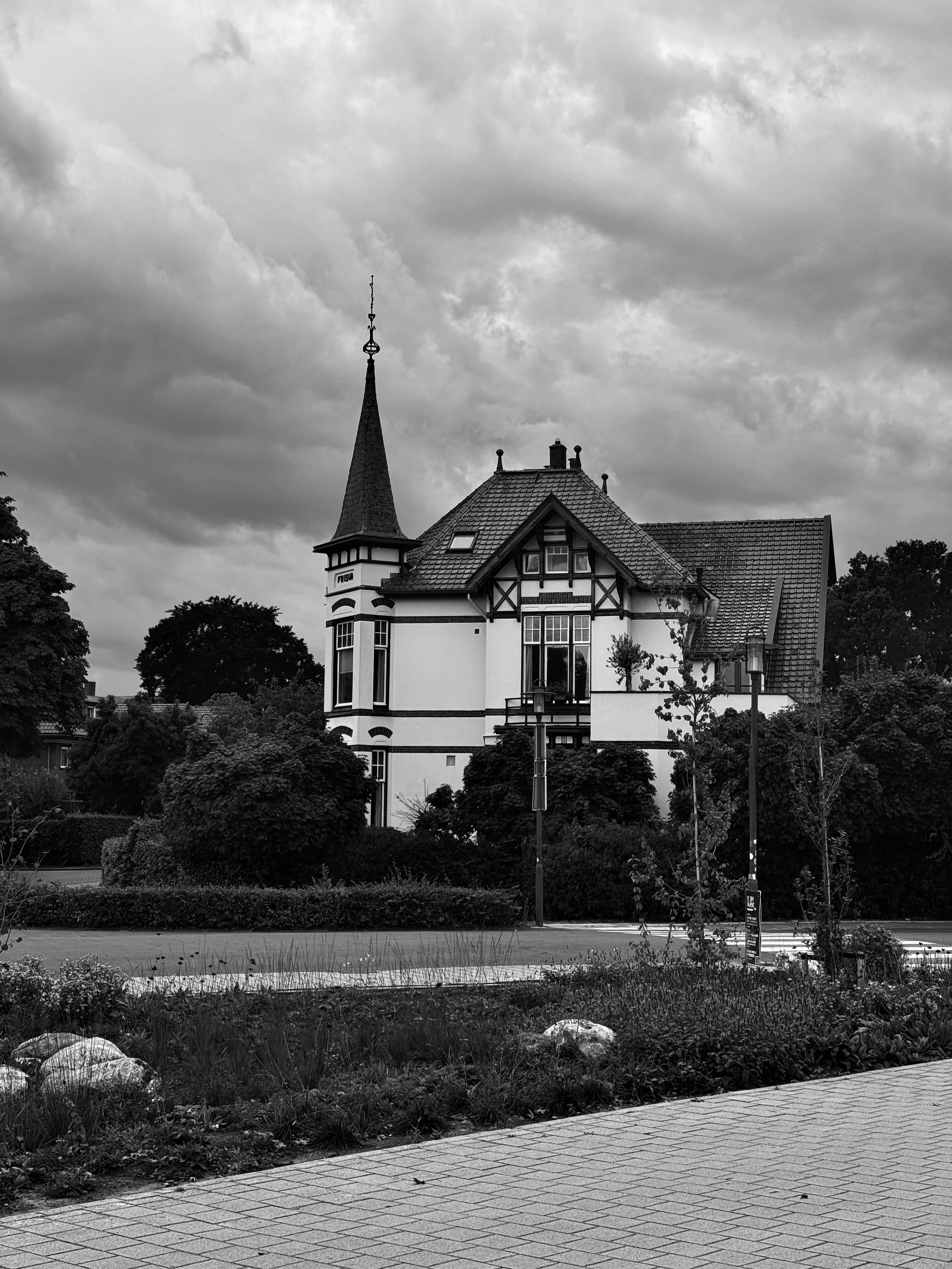 Black and white photo of a large house with a steeply pitched roof, tall spire, surrounded by trees and streetlamps.