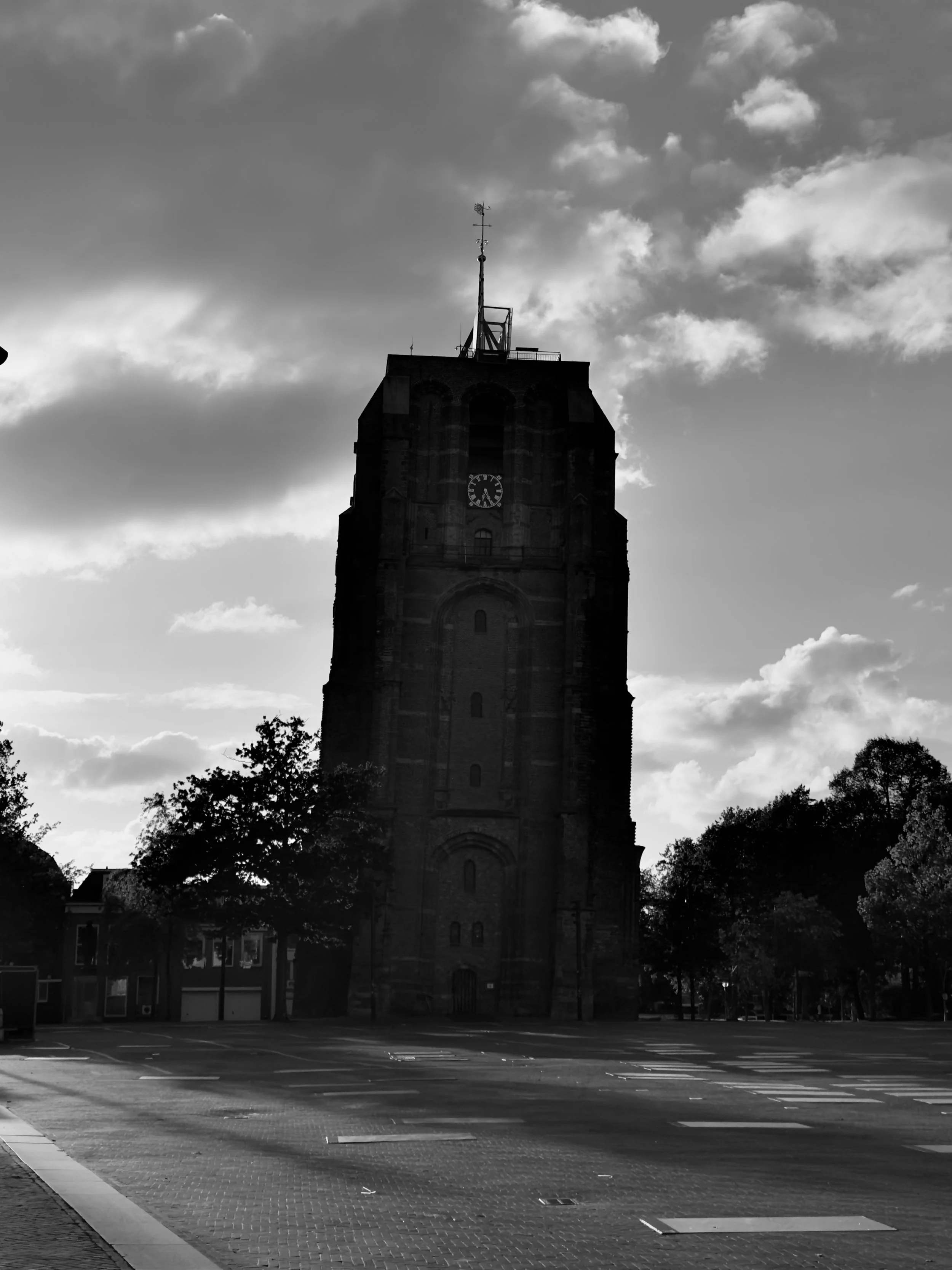 A large, historic clock tower with a weather vane on top, silhouetted against a partly cloudy sky, with an empty parking lot and trees in the foreground.