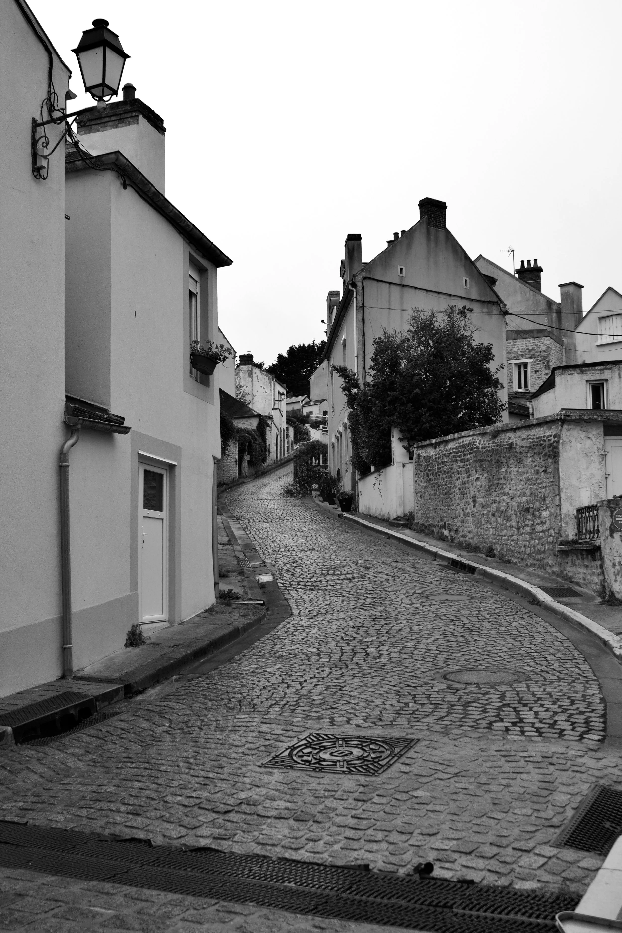 A black-and-white photo of a narrow cobblestone street winding uphill through a residential neighborhood with white houses and stone walls on either side.
