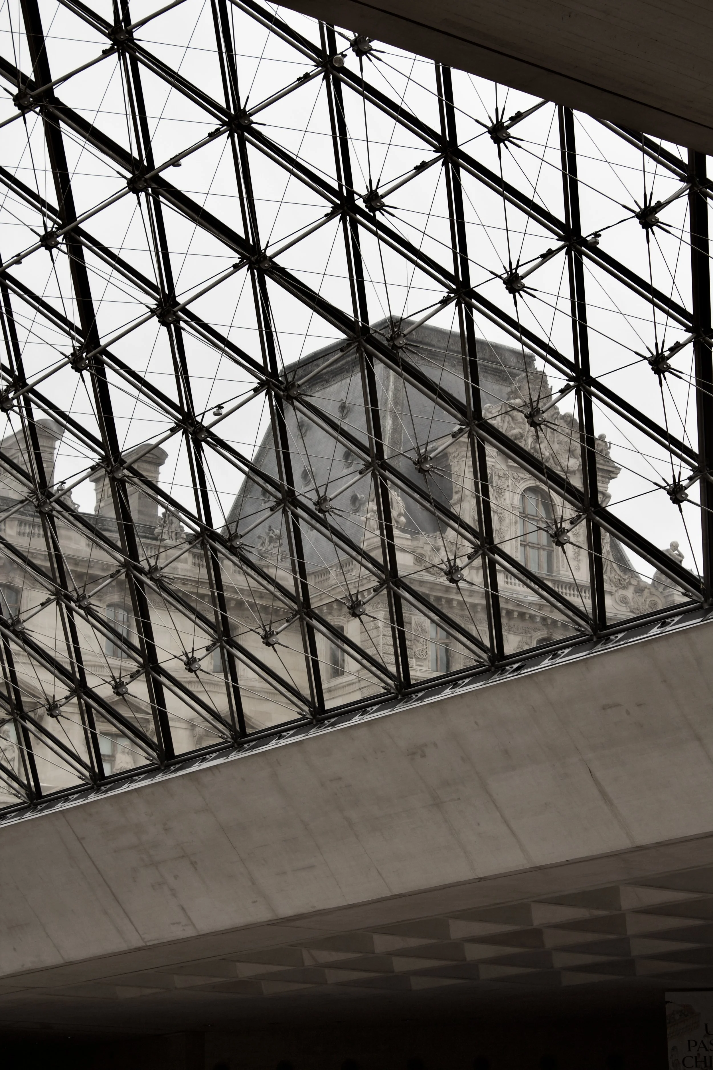 View of an ornate historic building through a glass and metal roof structure, with the building appearing in the background, overcast sky.